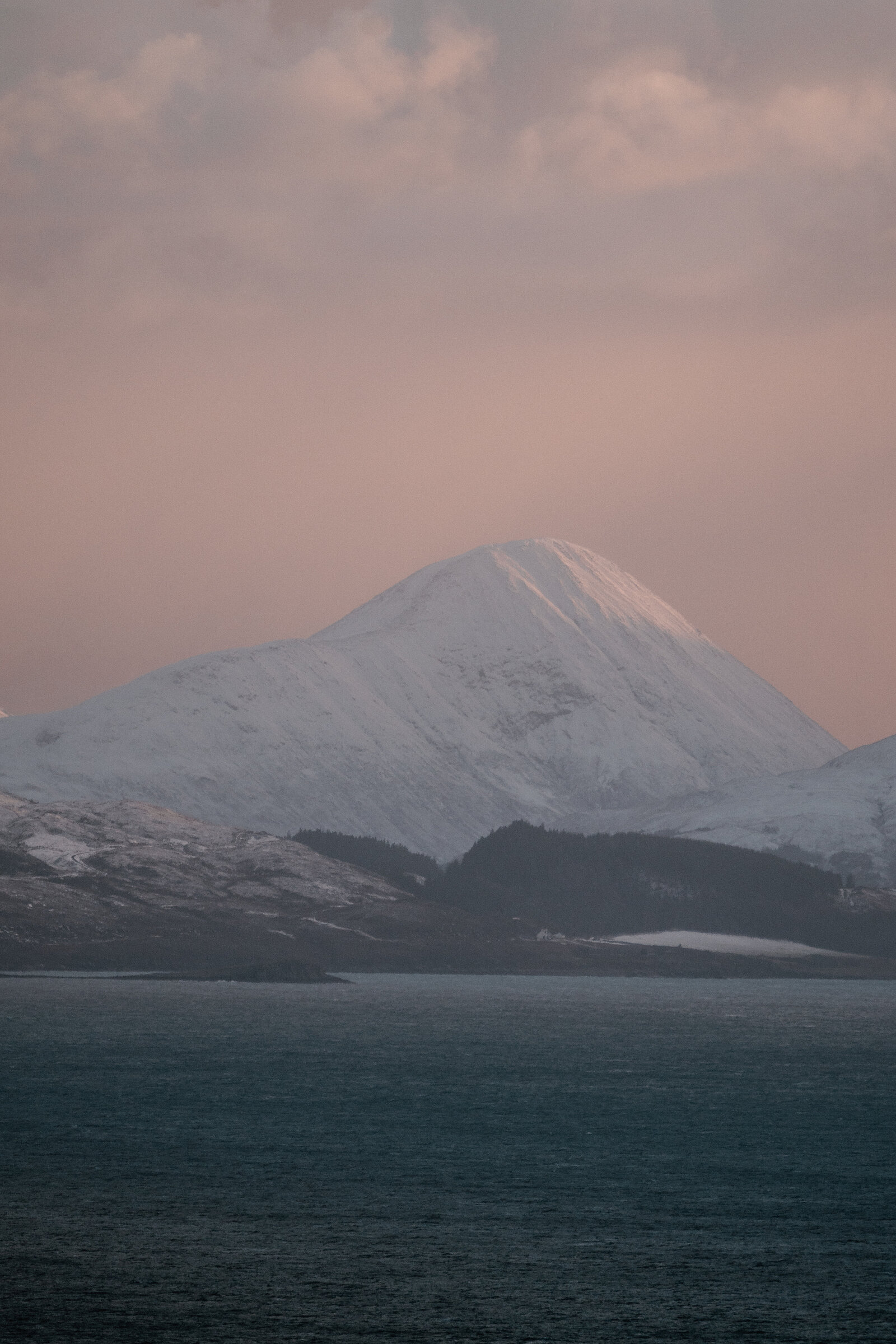 A single snow-capped peak stands above dark hillside forest beneath glowing evening cloud.