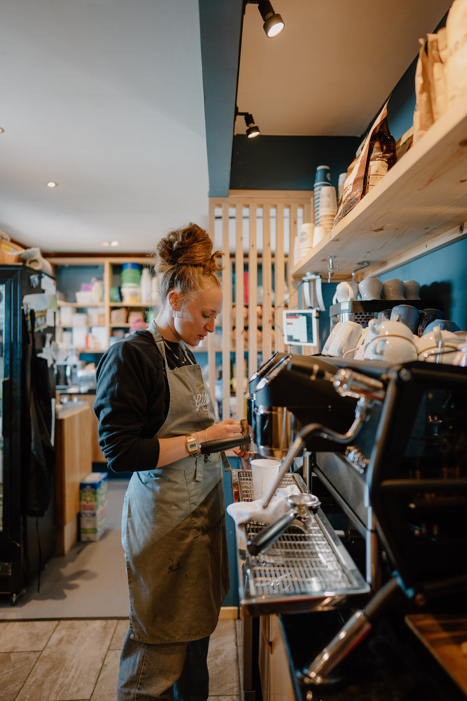 A barista in an apron works at an espresso machine in a shop with dark blue walls and wooden shelving.