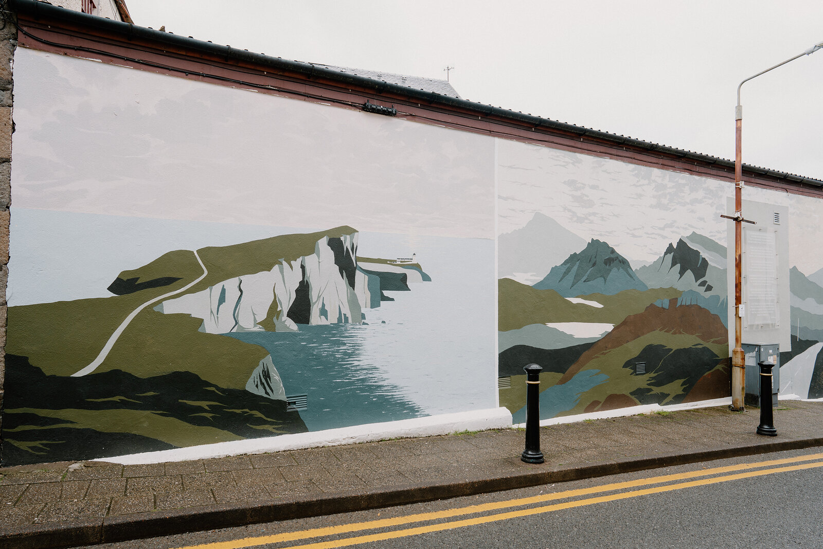A painted mural depicting a cliff-top path and jagged peaks stretches along a stone wall beside a pavement.
