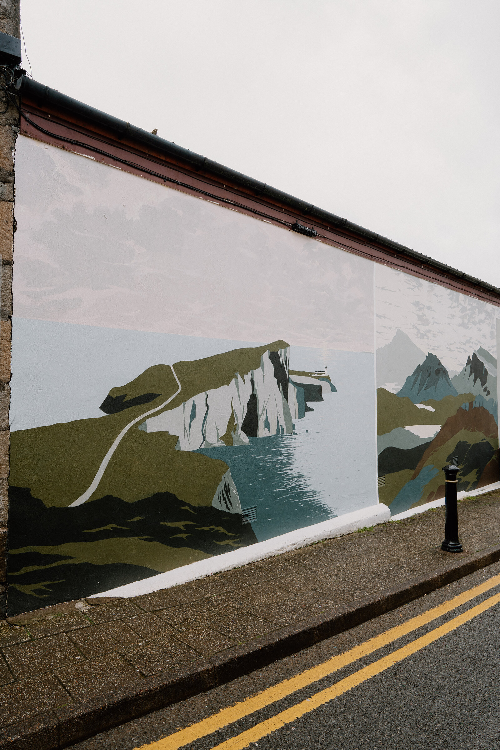 A colourful mural of a coastal headland and mountain range covers a long wall above a cobbled pavement.