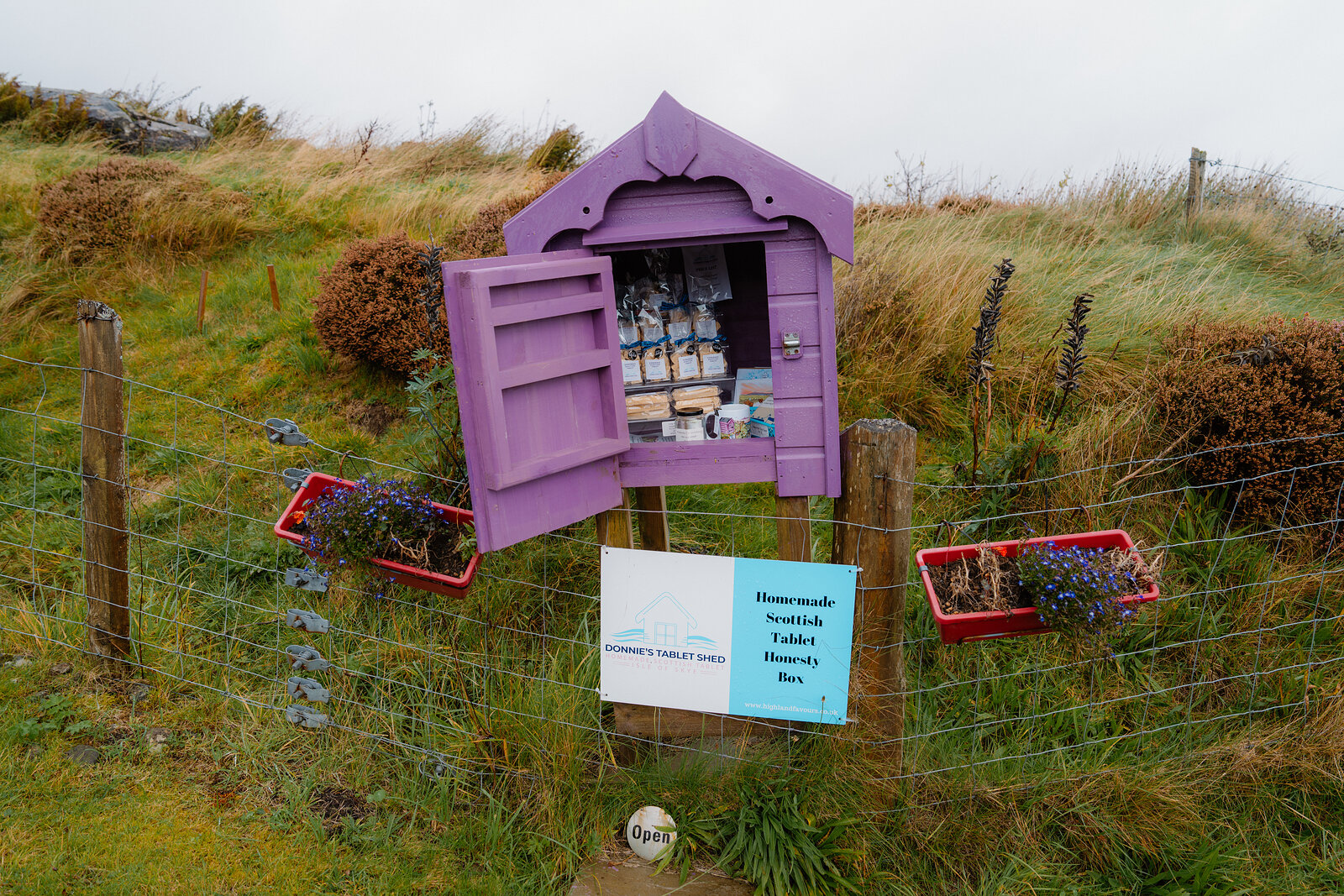 A purple wooden honesty box stocked with sweets stands open beside potted flowers and a fence.