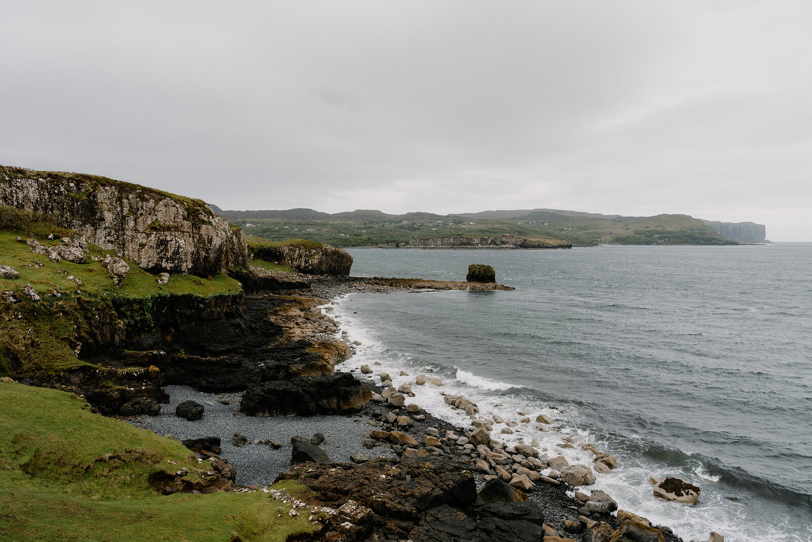 A rocky cove with a small sea stack sits beneath cliffs on the Isle of Skye, a distant headland beyond.
