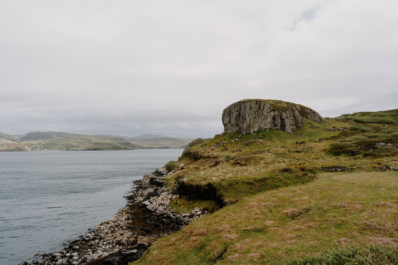 A rocky outcrop rises above a grassy headland on the Isle of Skye, a sea loch beyond.