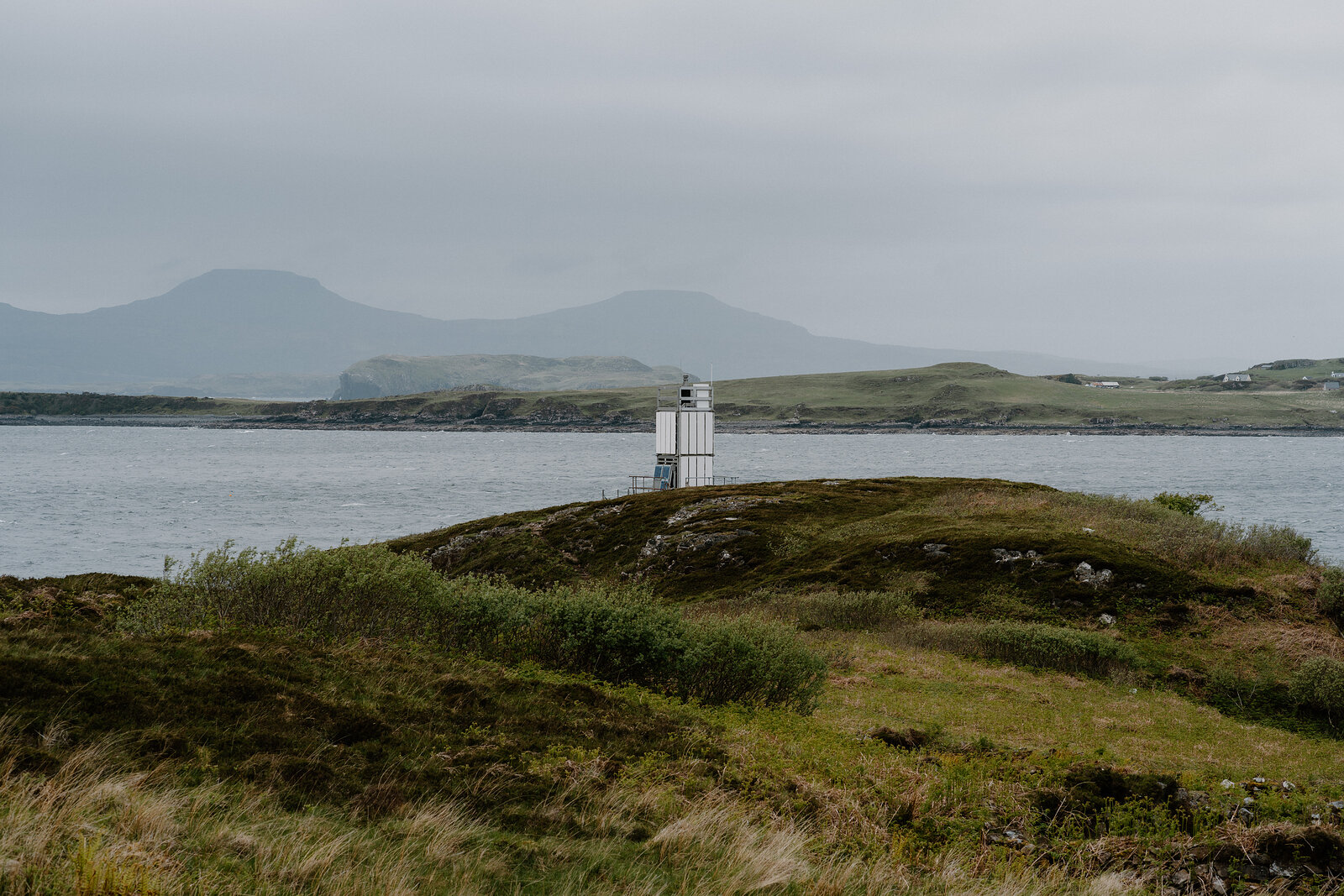 A white beacon tower stands on a grassy point above the water, hills visible across the strait on the Isle of Skye.