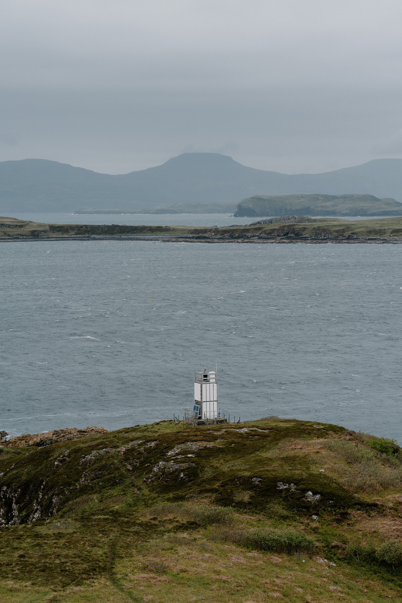 A white beacon tower stands on a grassy point overlooking a strait, hills rising beyond on the Isle of Skye.
