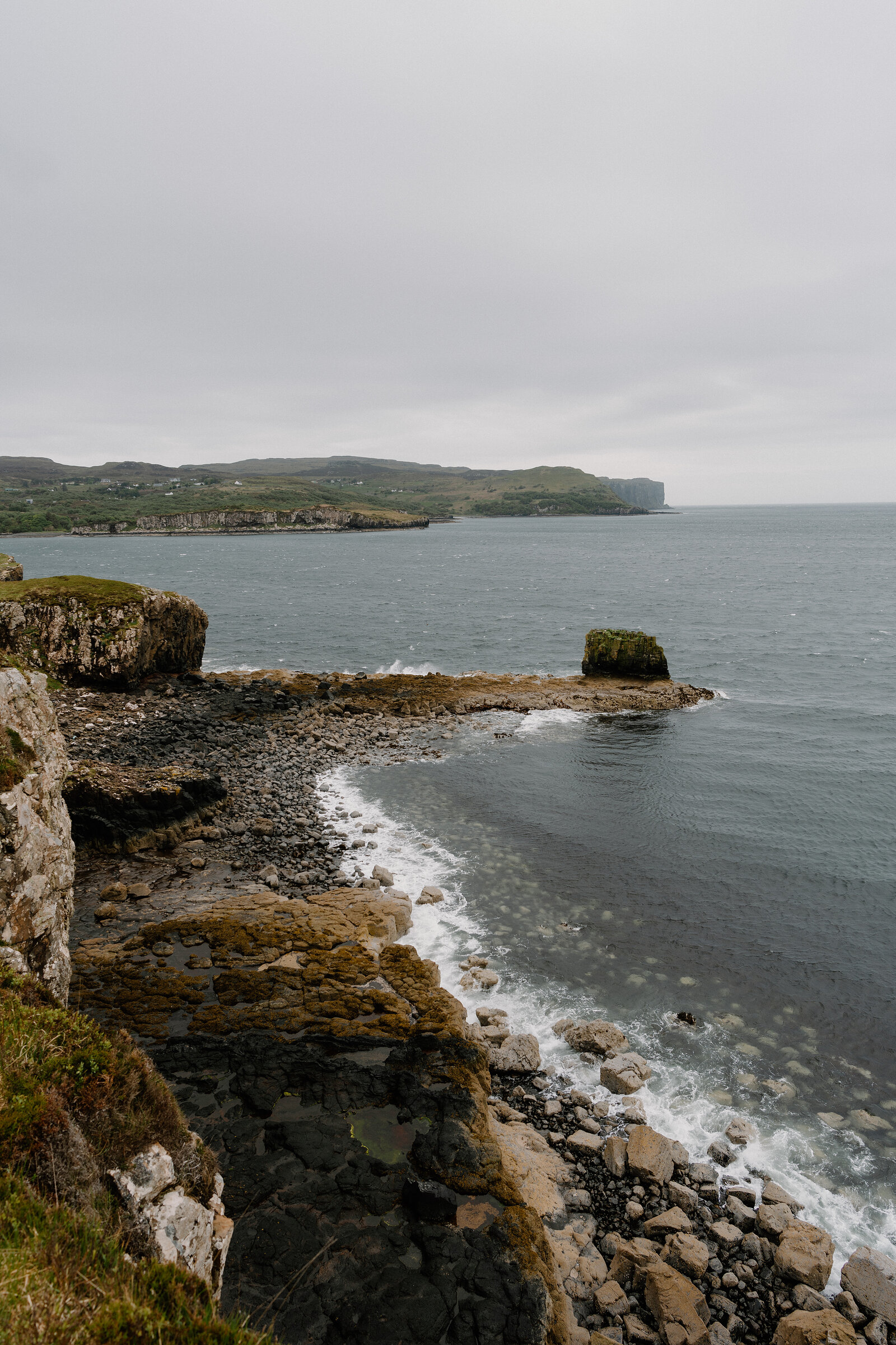 A small grassy sea stack sits offshore in a rocky bay on the Isle of Skye.