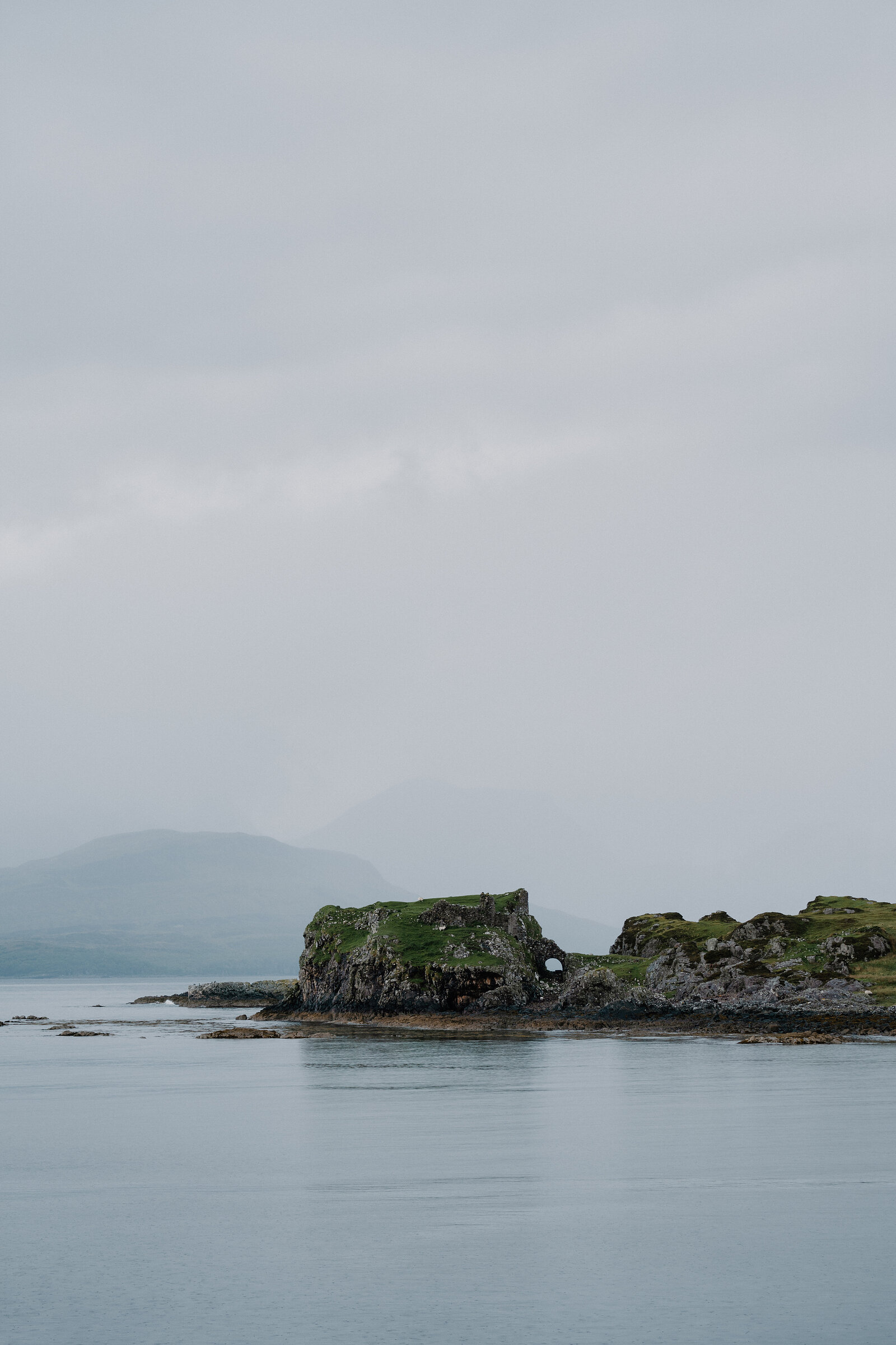 A ruined stone castle sits on a small tidal islet across calm loch water on the Isle of Skye.