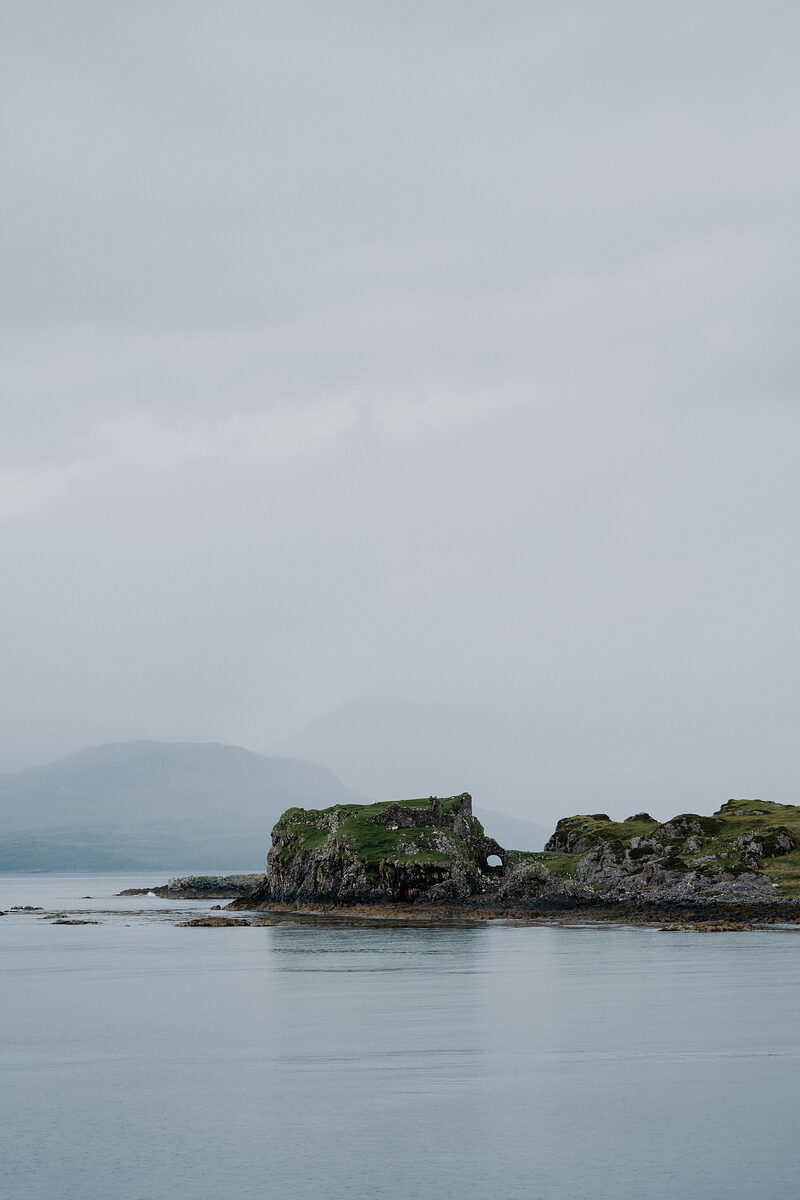 A ruined stone castle sits on a small tidal islet across calm loch water on the Isle of Skye.