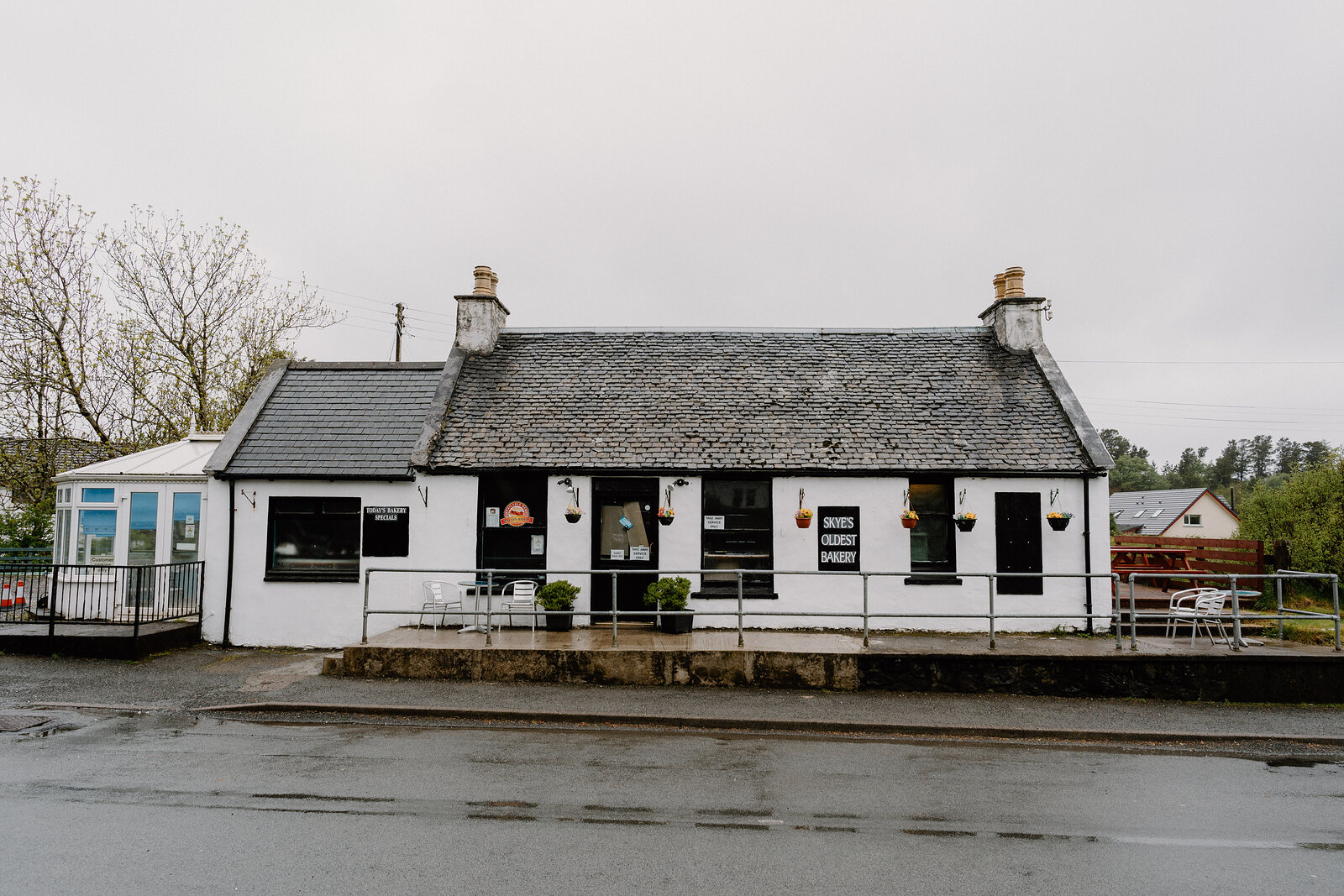 Skye's Oldest Bakery's whitewashed cottage sits beside a rain-slicked road, with outdoor tables and hanging flower baskets at its door.
