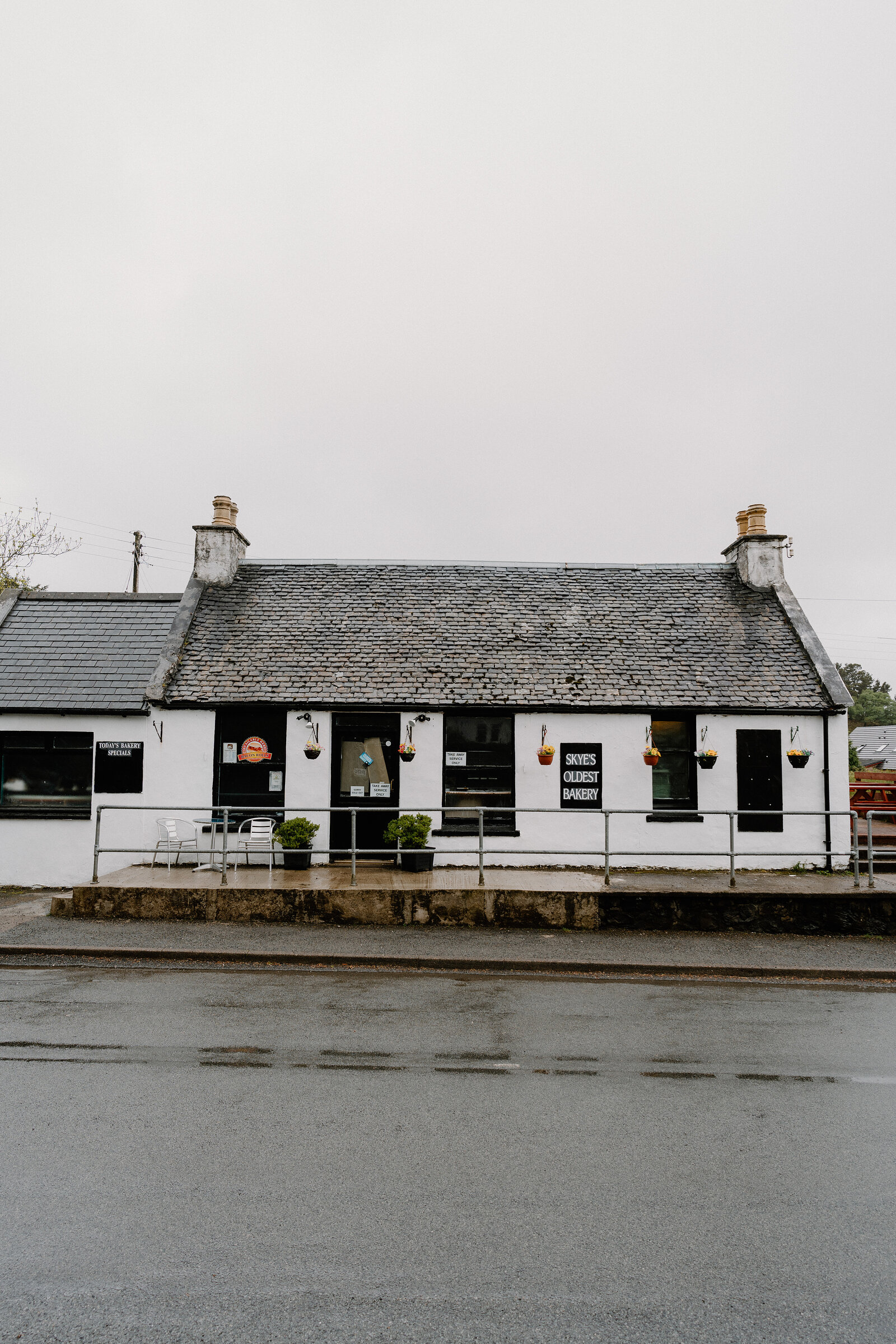 Skye's Oldest Bakery's cottage exterior shows its sign among hanging flower baskets and chimney pots under an overcast sky.