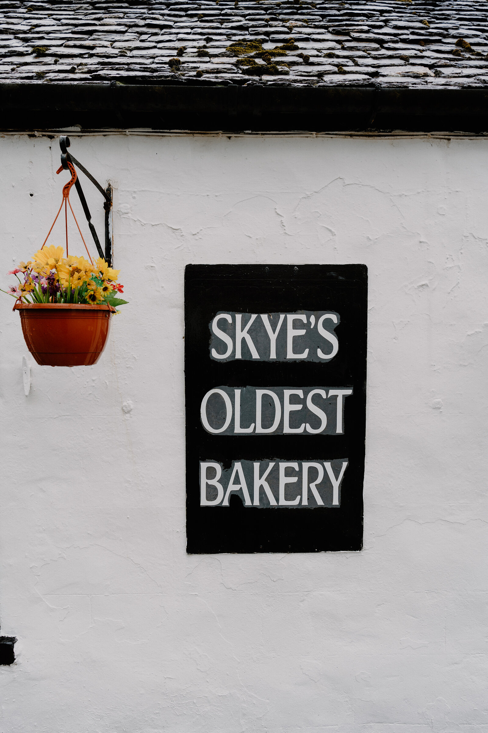 A hanging basket of yellow flowers hangs beside the black-and-white 'SKYE'S OLDEST BAKERY' sign on a whitewashed wall.