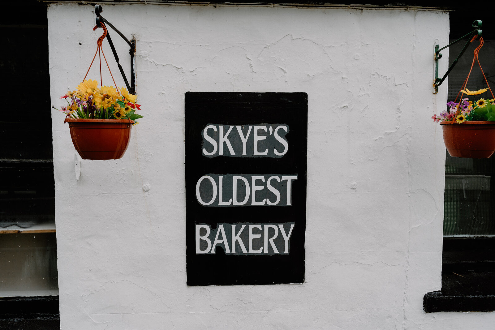 Two hanging baskets of flowers flank the black-and-white 'SKYE'S OLDEST BAKERY' sign mounted on a whitewashed wall.