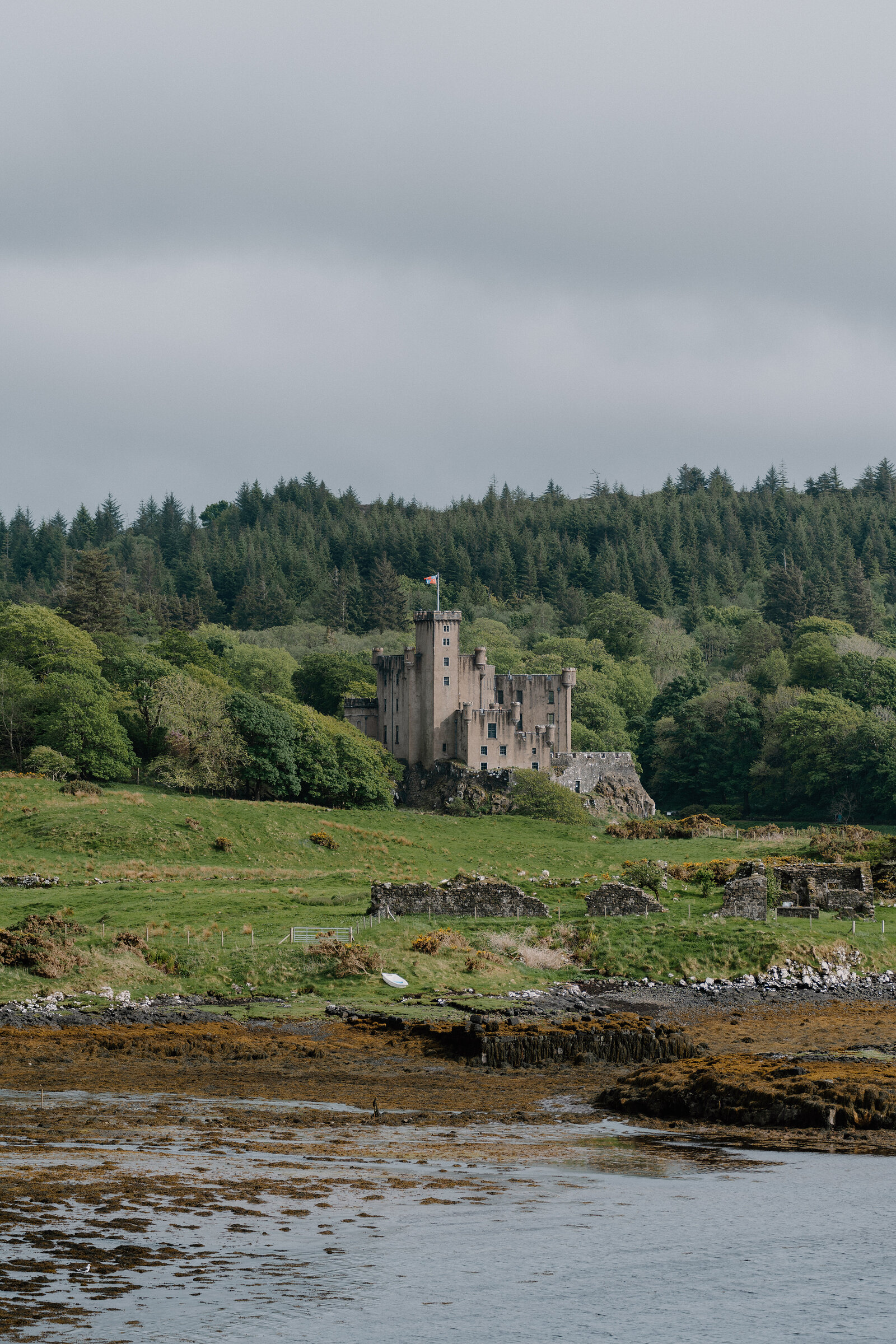 Dunvegan Castle's turreted keep rises above trees on the shore of a sea loch, with seaweed-strewn rocks in the foreground.
