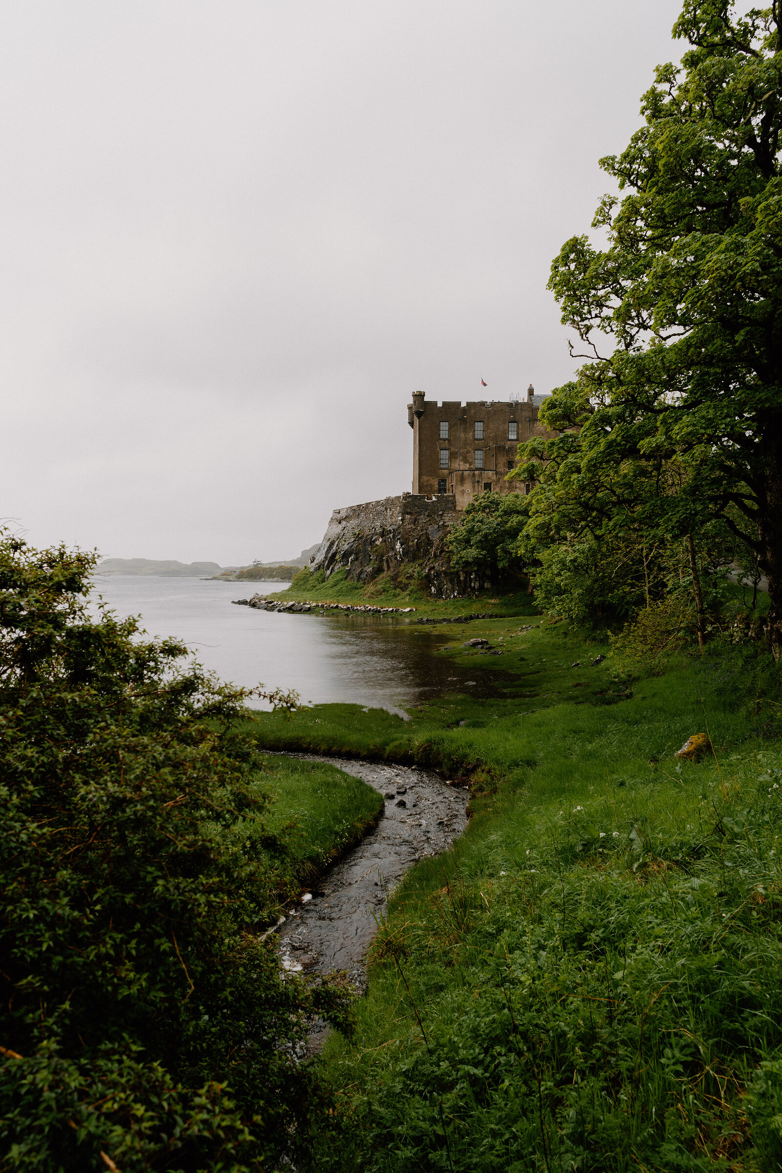 Dunvegan Castle stands on a rocky point above a sea loch, with a stream winding through grass in the foreground.