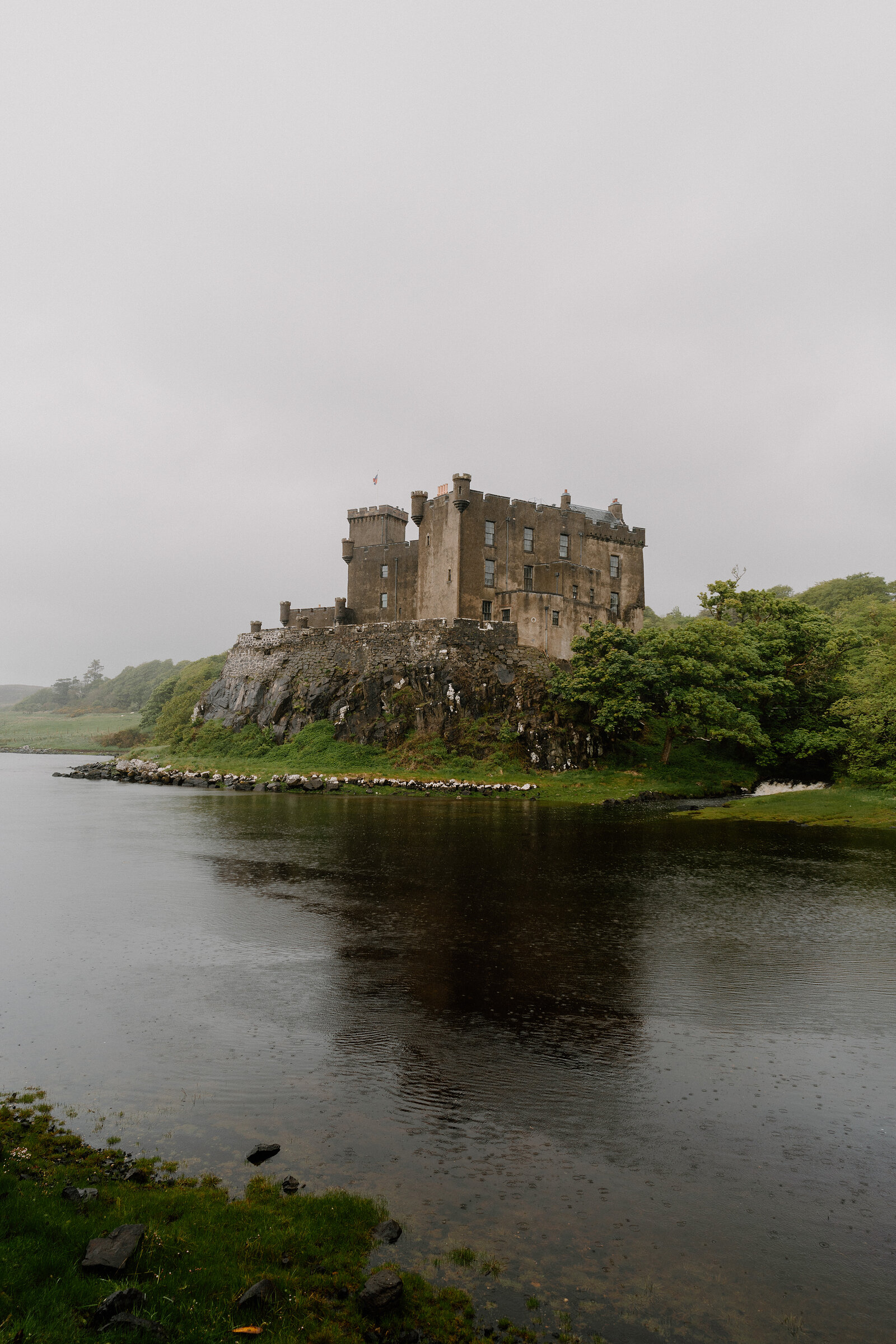 Dunvegan Castle rises above wooded shoreline, reflected in the still water of the loch.