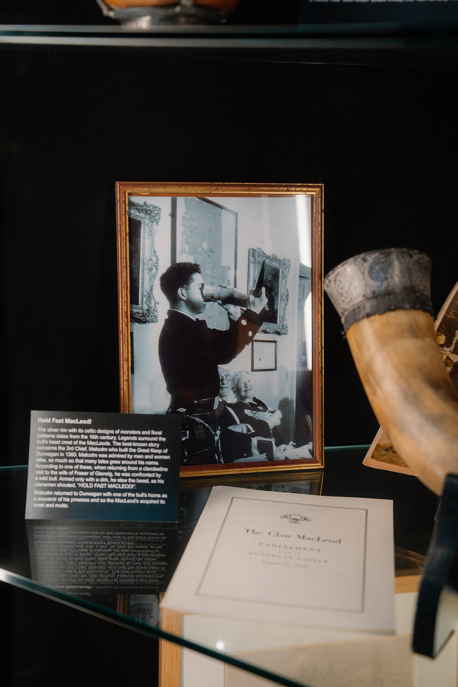 A framed photograph captioned 'Hold Fast MacLeod!' stands beside a horn and a Clan MacLeod parliament programme at Dunvegan Castle.
