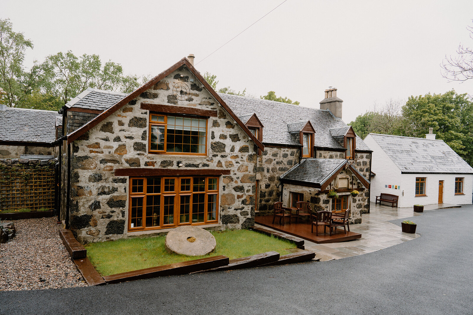 A stone cottage with a wooden deck and outbuildings sits beside a gravel driveway.