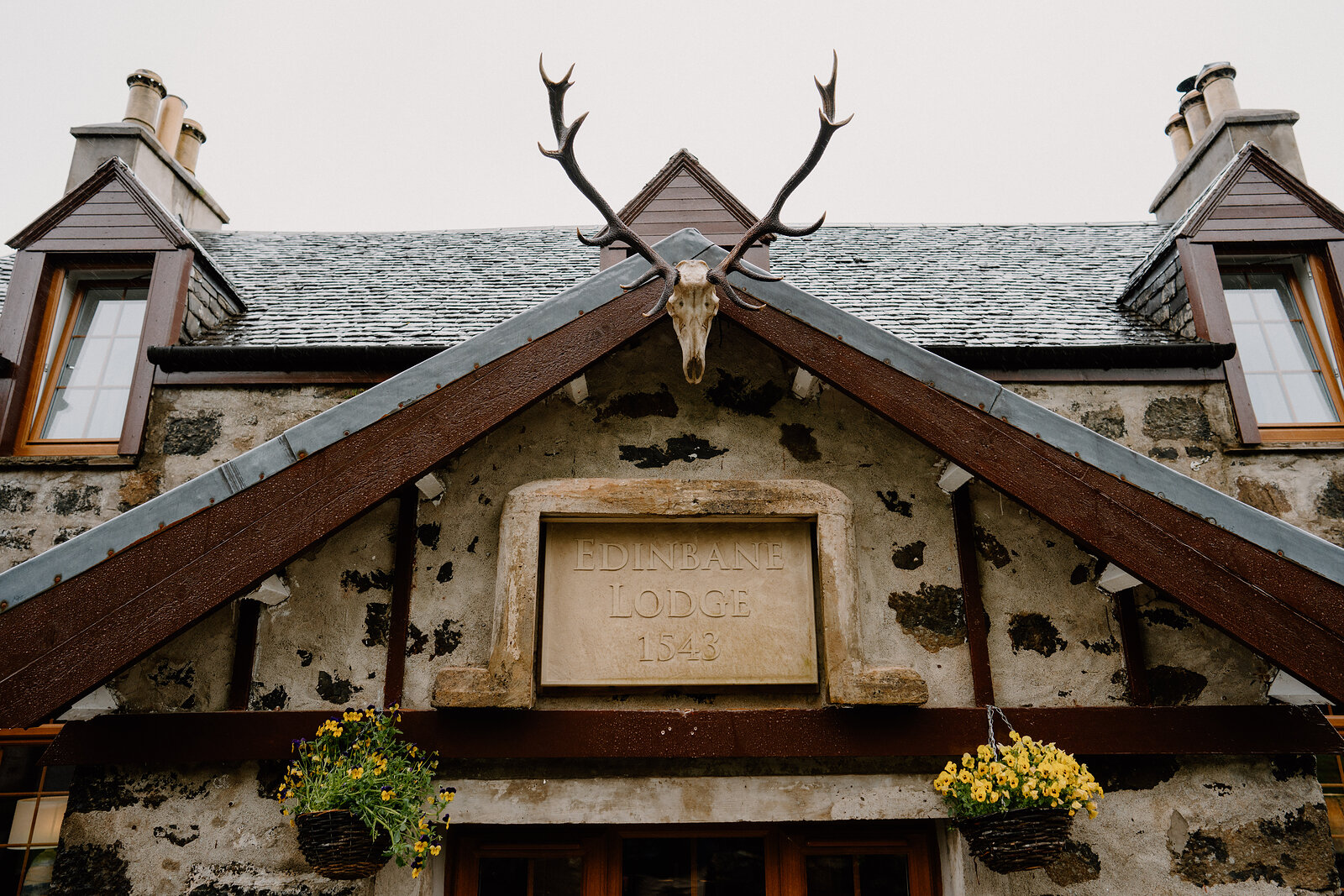 A stag skull with antlers is mounted above Edinbane Lodge's gabled stone entrance, its datestone flanked by hanging flower baskets.