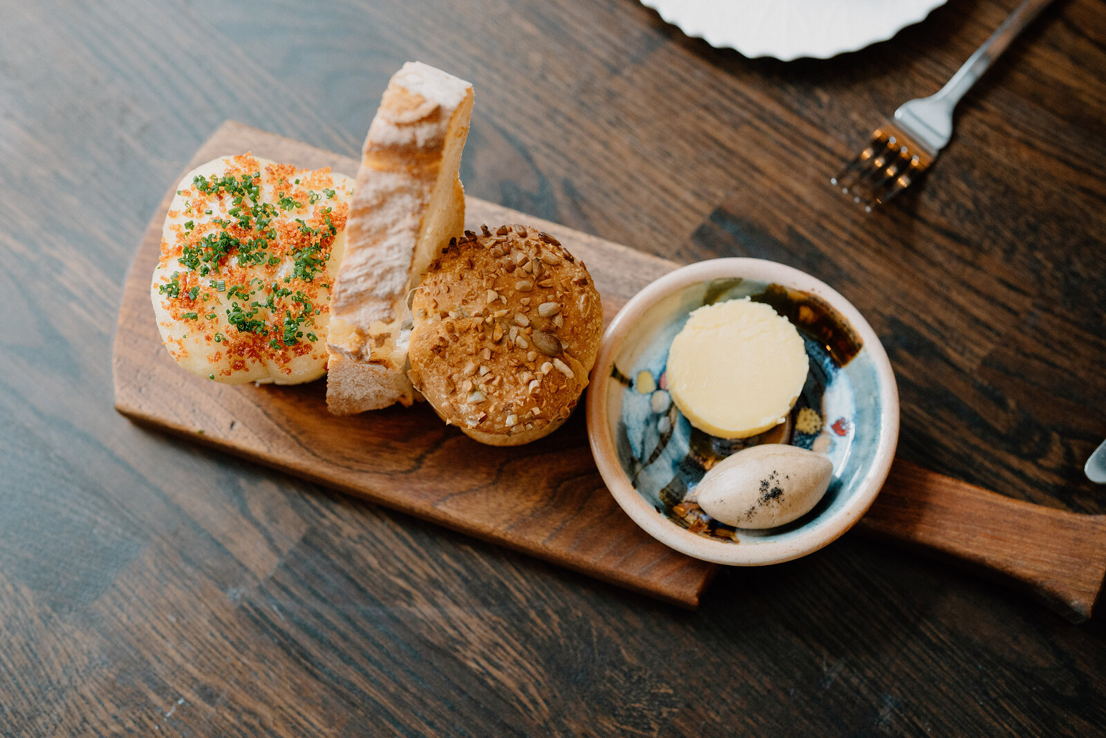 Bread and a roll are arranged beside whipped butter and a bulb of garlic on a wooden board.
