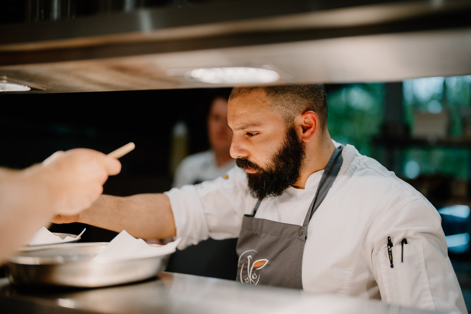 A chef gestures while plating a dish beside a colleague at a stainless steel pass at Edinbane Lodge.