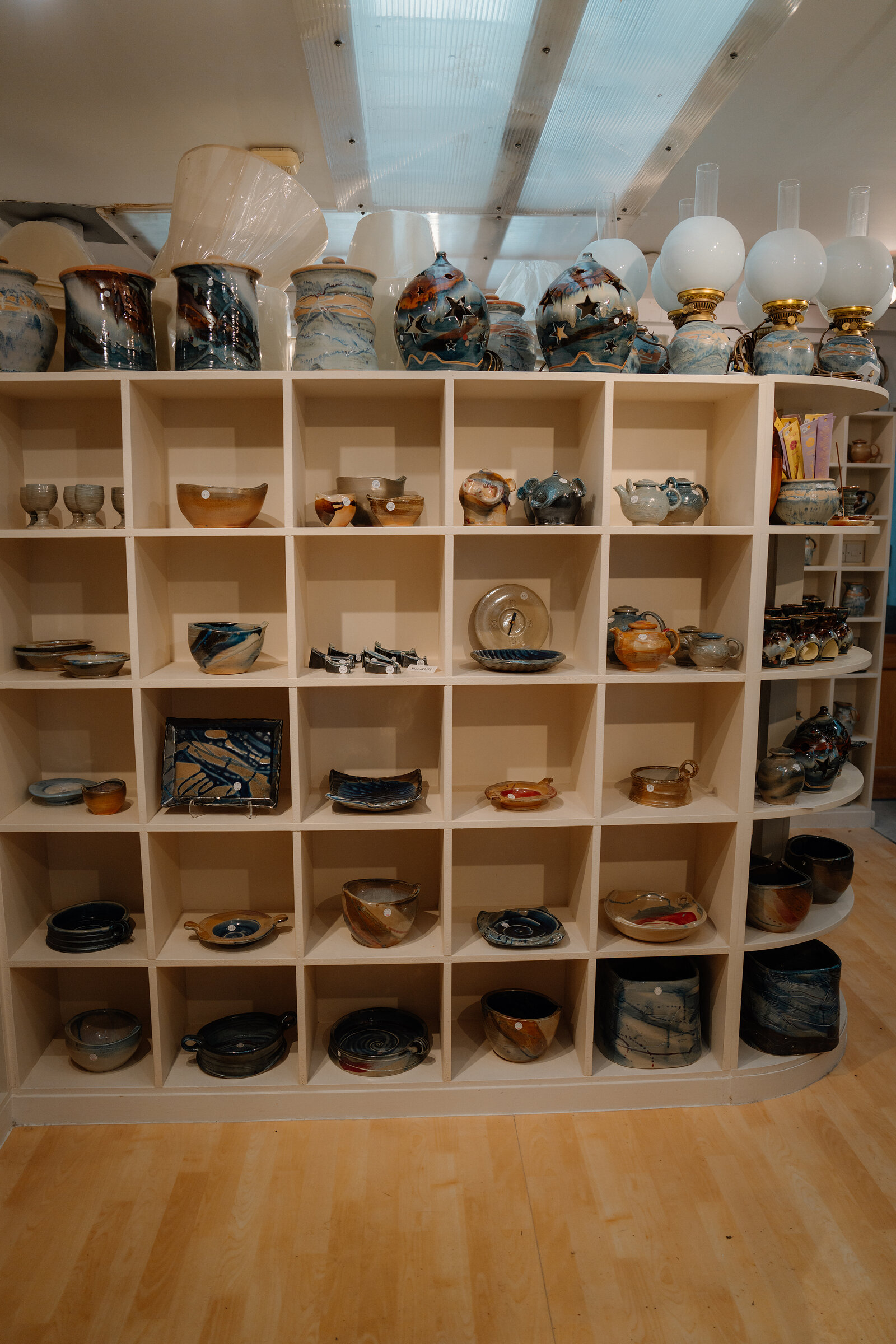 Shelves of hand-glazed stoneware bowls, plates and vases fill a pottery studio beneath skylights and glass lampshades.