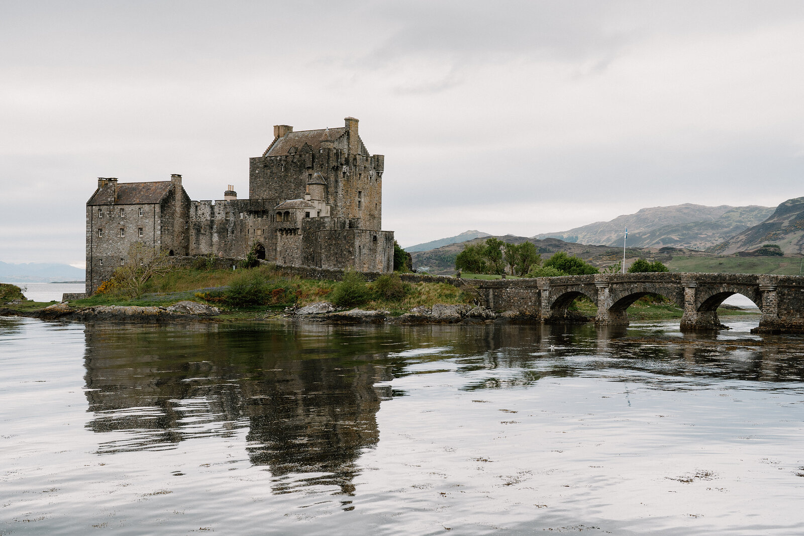Eilean Donan Castle and its stone footbridge are reflected in the calm loch beneath hills.