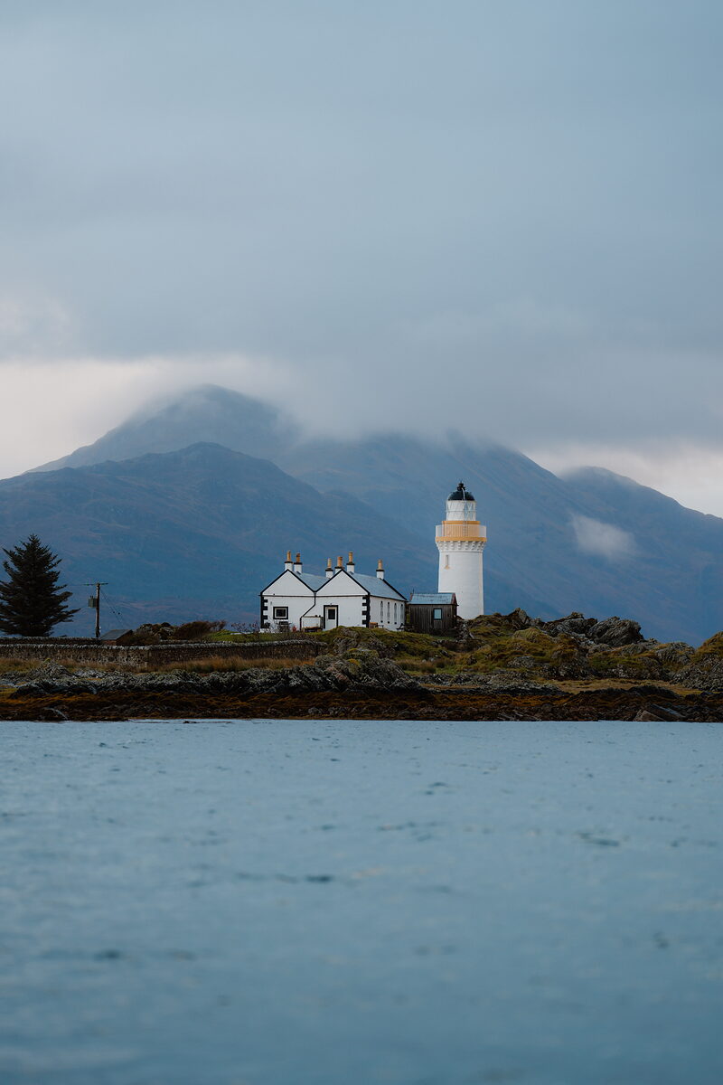 A white lighthouse and keeper's cottages stand on a rocky islet beneath misty hills.