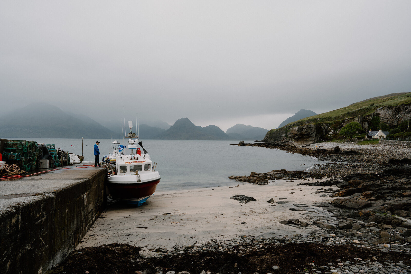 A fishing boat is moored at a stone slipway with misty mountains rising behind on the Isle of Skye.