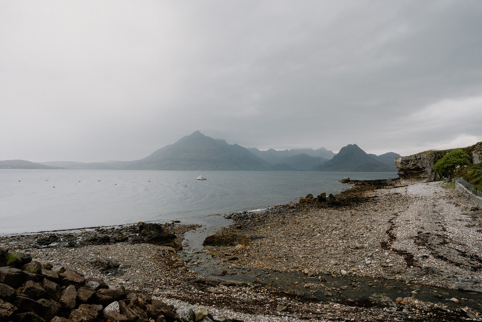 A pebble shoreline curves toward a misty bay with mountains rising beyond on the Isle of Skye.