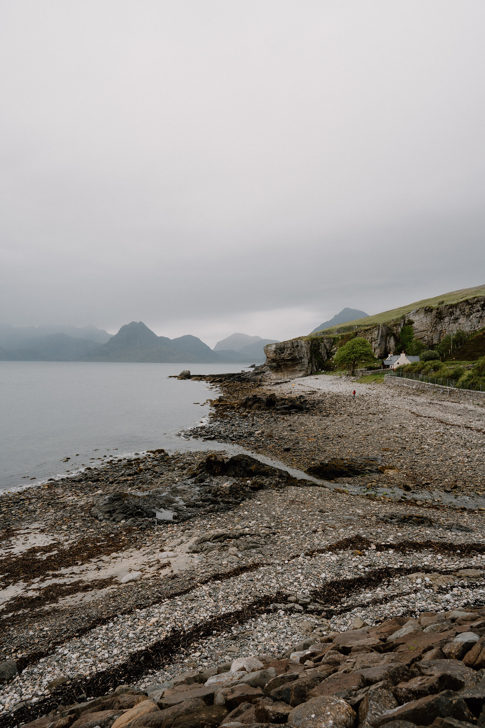 A pebble beach curves beneath misty mountains, with a cottage perched on the cliff edge on the Isle of Skye.