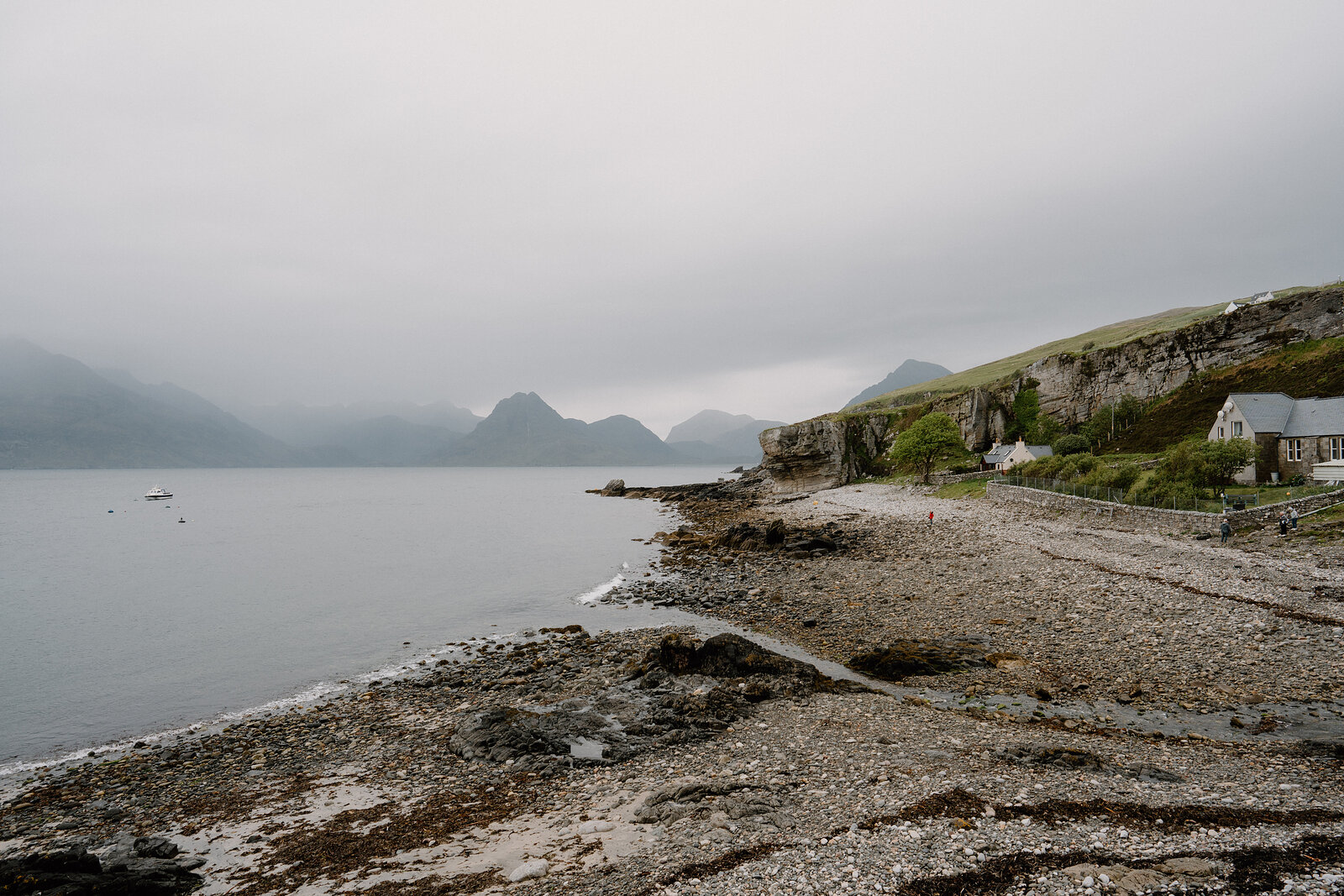 Calm water meets a pebble shore below misty mountains and a cliffside cottage on the Isle of Skye.