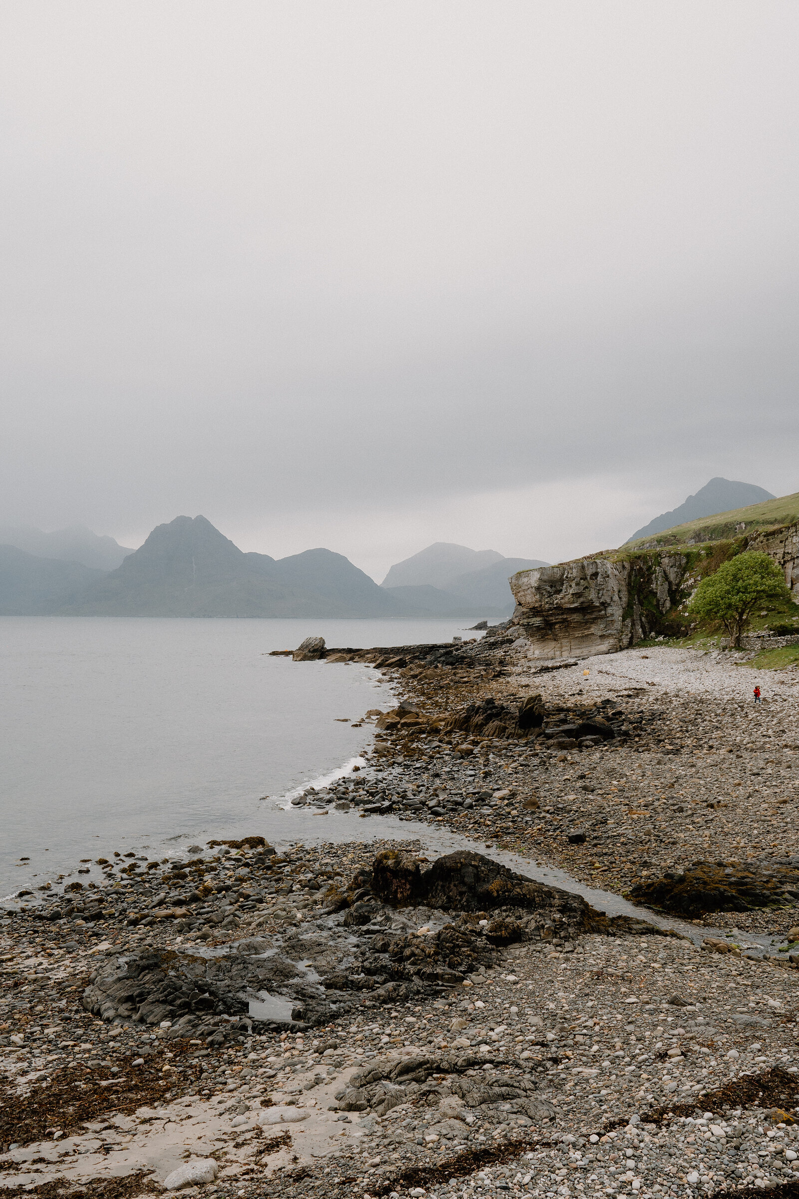 A figure in red walks along a pebble beach beneath misty mountains on the Isle of Skye.
