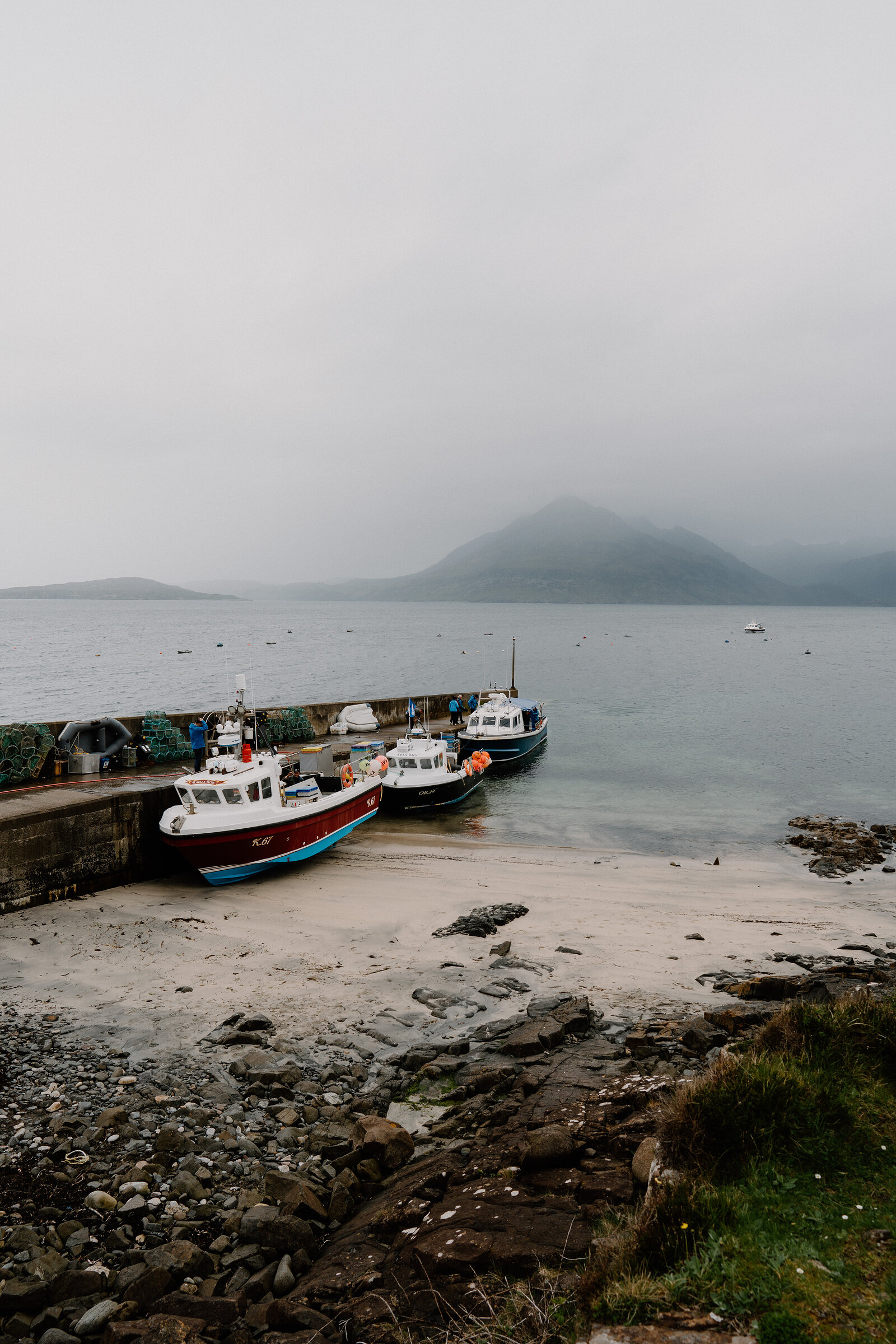Fishing boats rest on the sand beside a stone slipway beneath a misty hillside on the Isle of Skye.