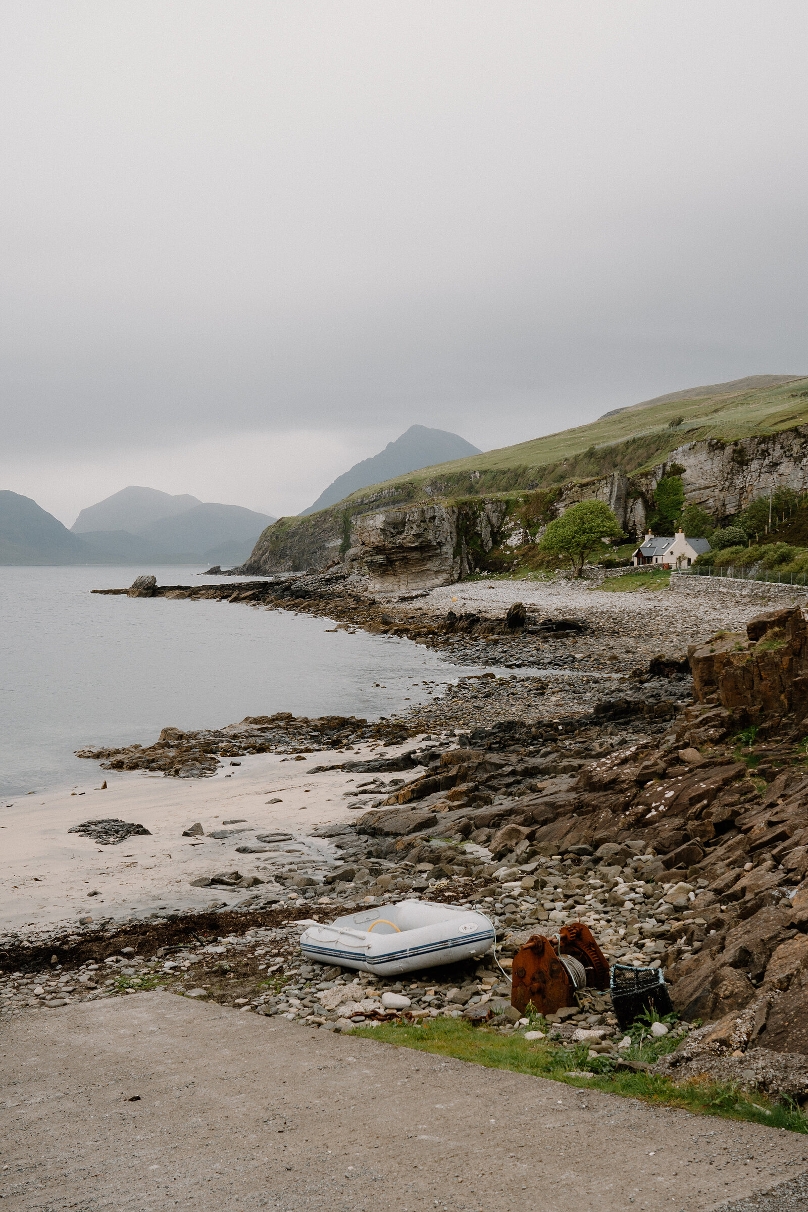 A white inflatable dinghy sits on rocks beside a pebble shore beneath cliffs and a distant cottage on the Isle of Skye.