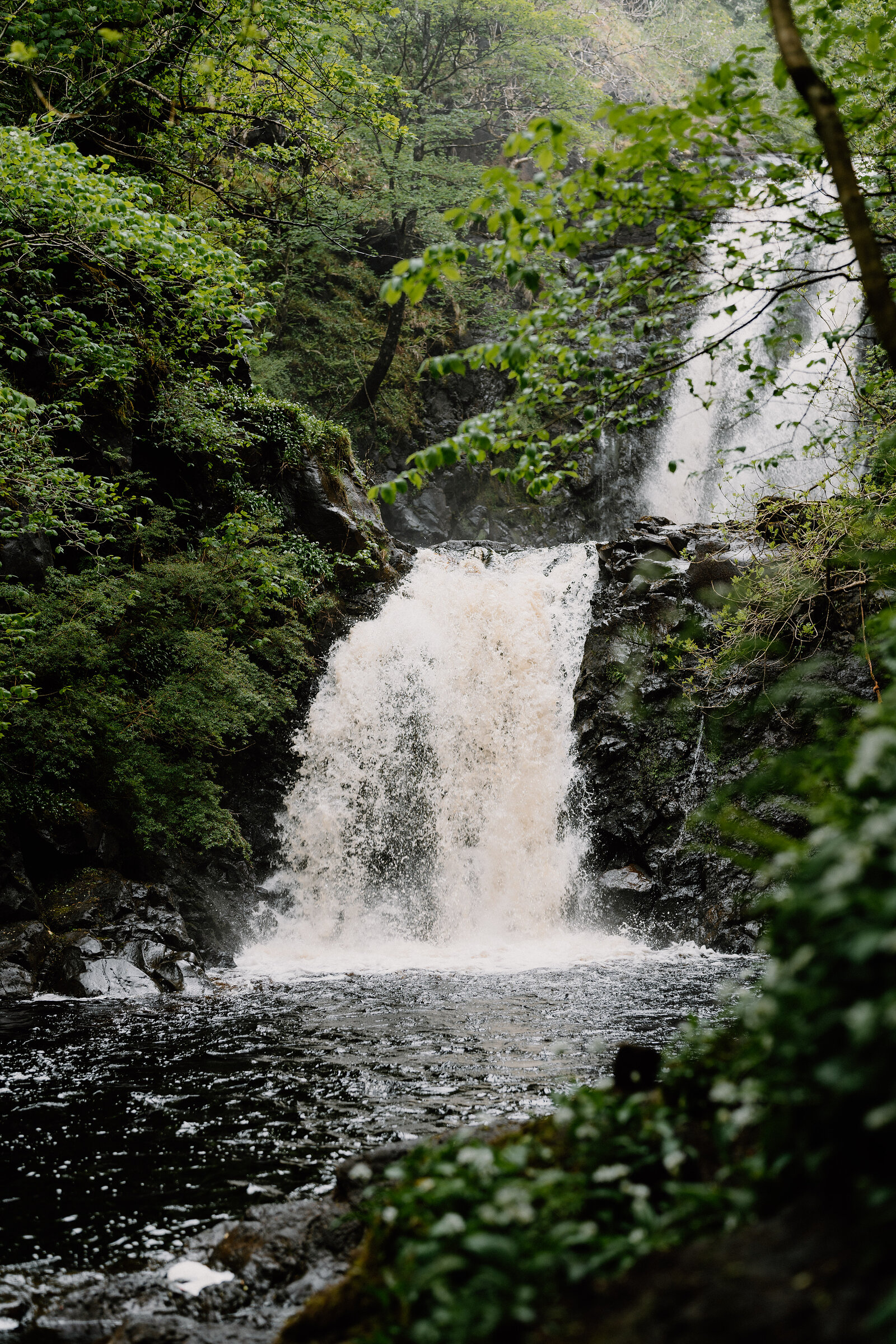 A two-tiered waterfall crashes into a pool in a green wooded gorge on the Isle of Skye.