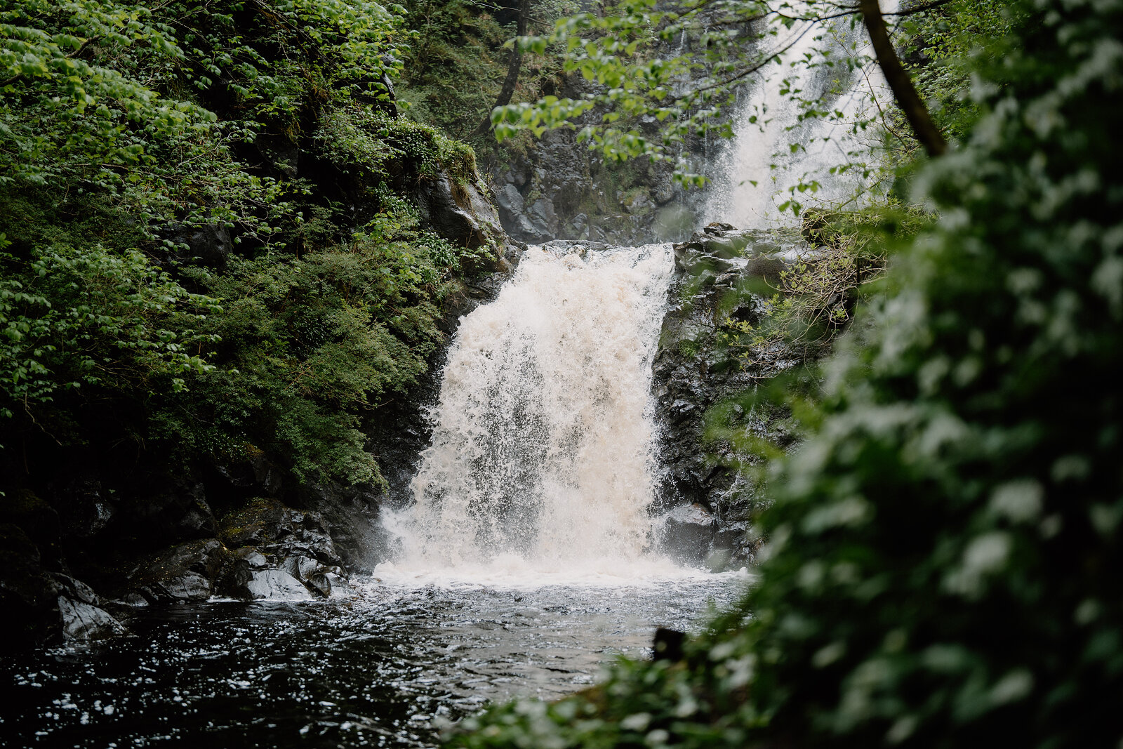 A waterfall drops through a narrow rock channel into a pool surrounded by dense woodland on the Isle of Skye.