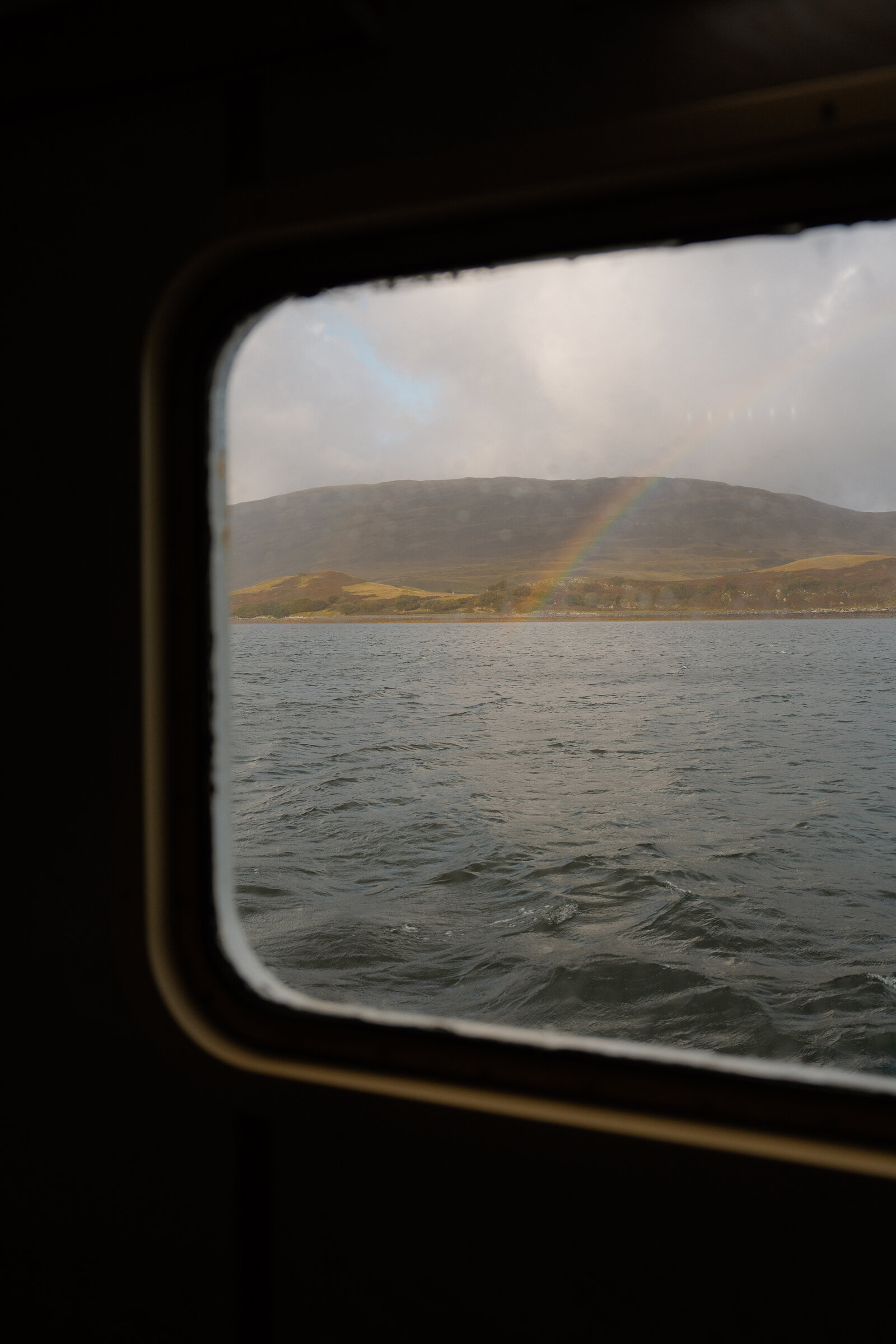 A rainbow arcs over hills viewed through a ferry window near the Isle of Skye.