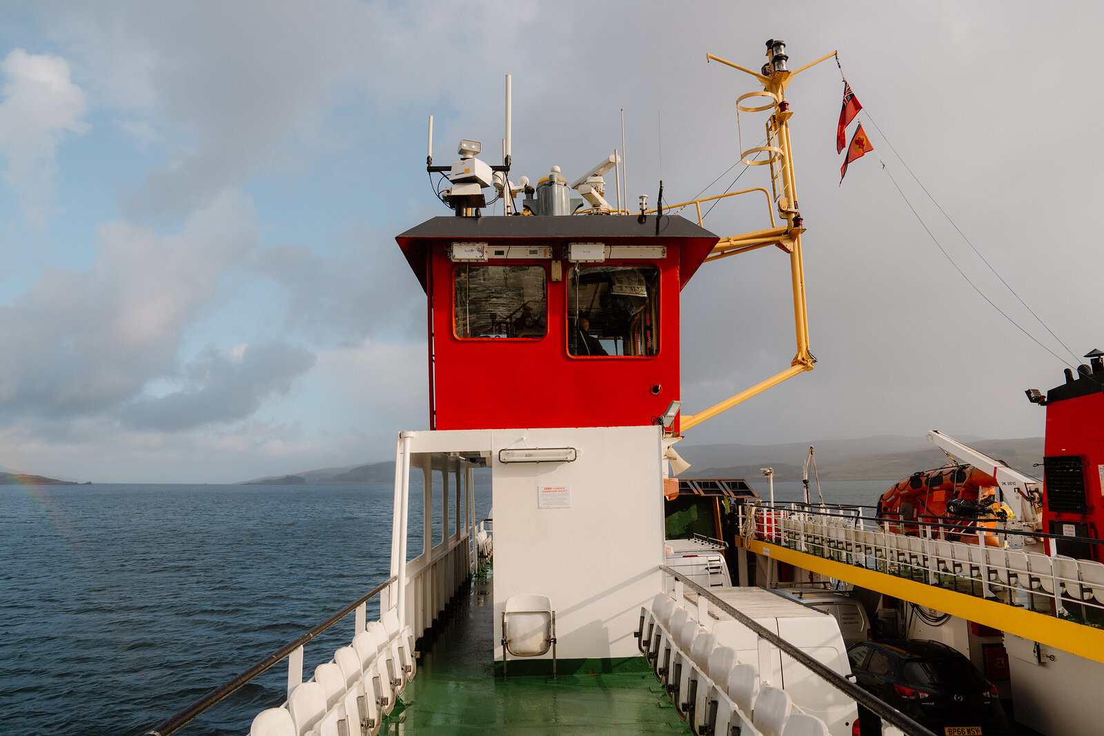 A ferry's red wheelhouse and radar mast rise above the deck as it crosses toward the Isle of Skye.