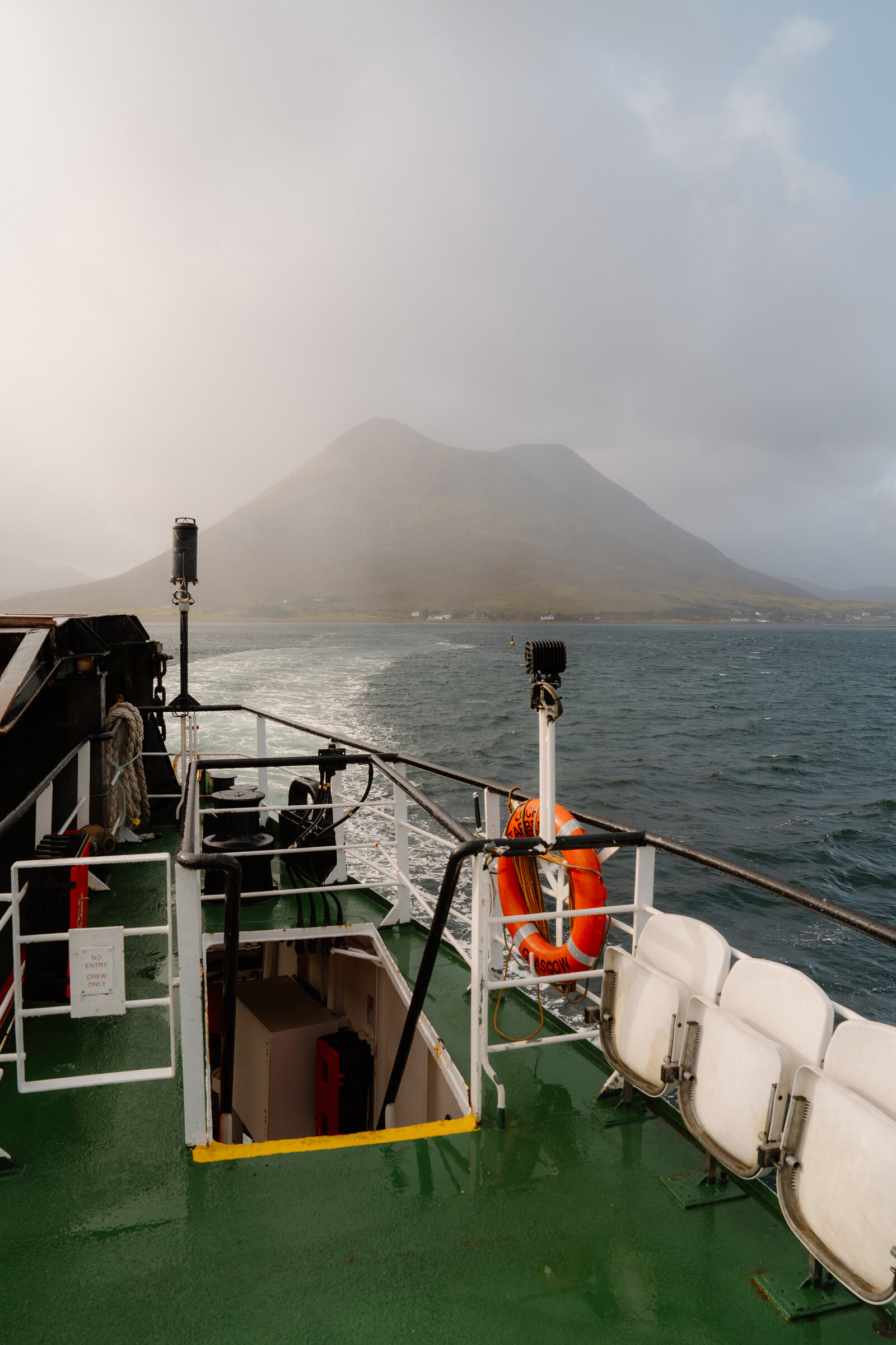 Mist shrouds a mountain beyond a ferry's wake, lifebelt and folded seating near the Isle of Skye.