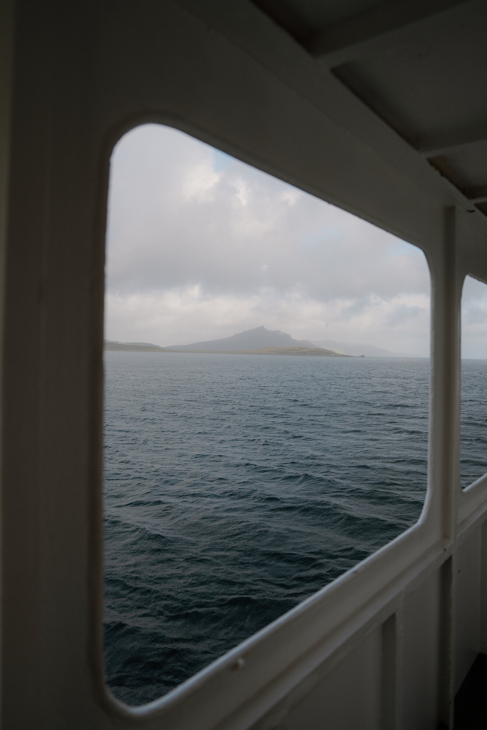A distant hill is framed by a ferry's window over calm water near the Isle of Skye.