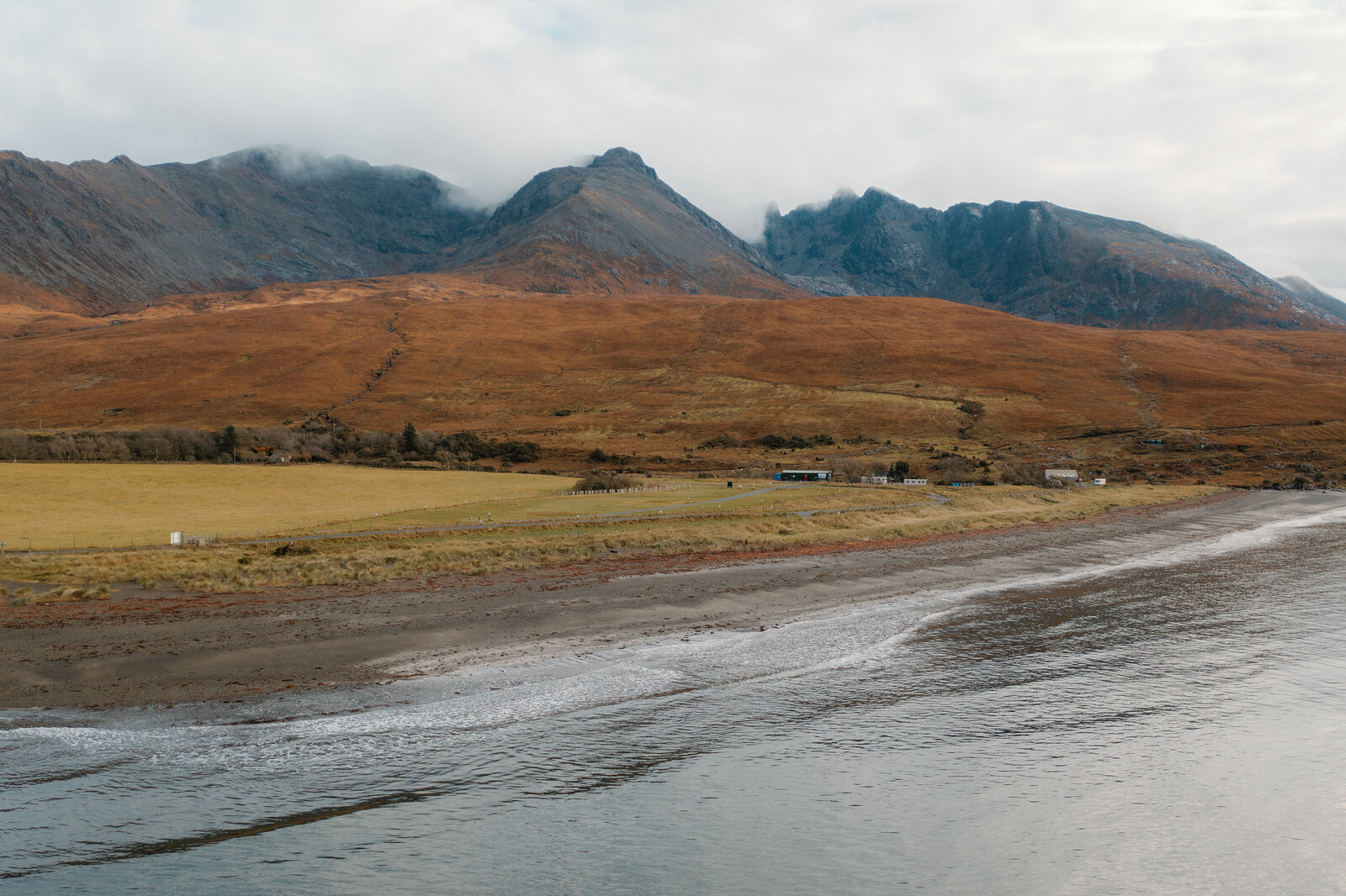 Autumn moorland curves around a sandy bay beneath the misty Cuillin mountains on the Isle of Skye.