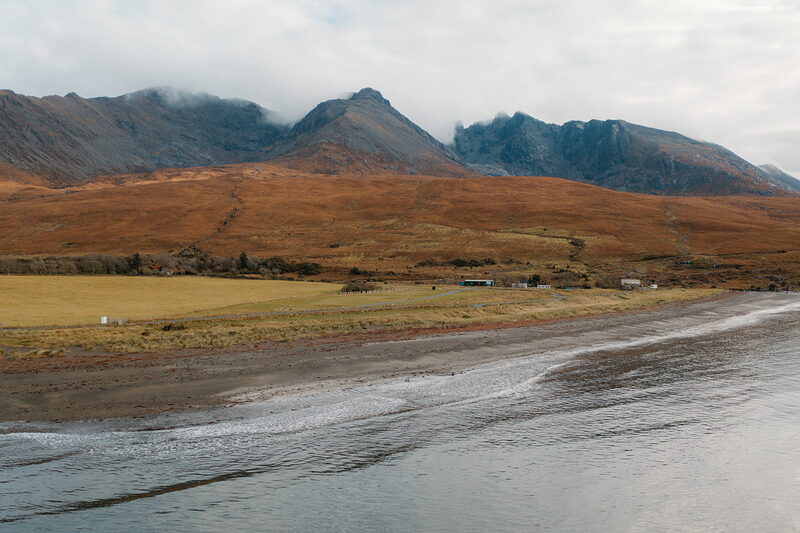 Autumn moorland curves around a sandy bay beneath the misty Cuillin mountains on the Isle of Skye.