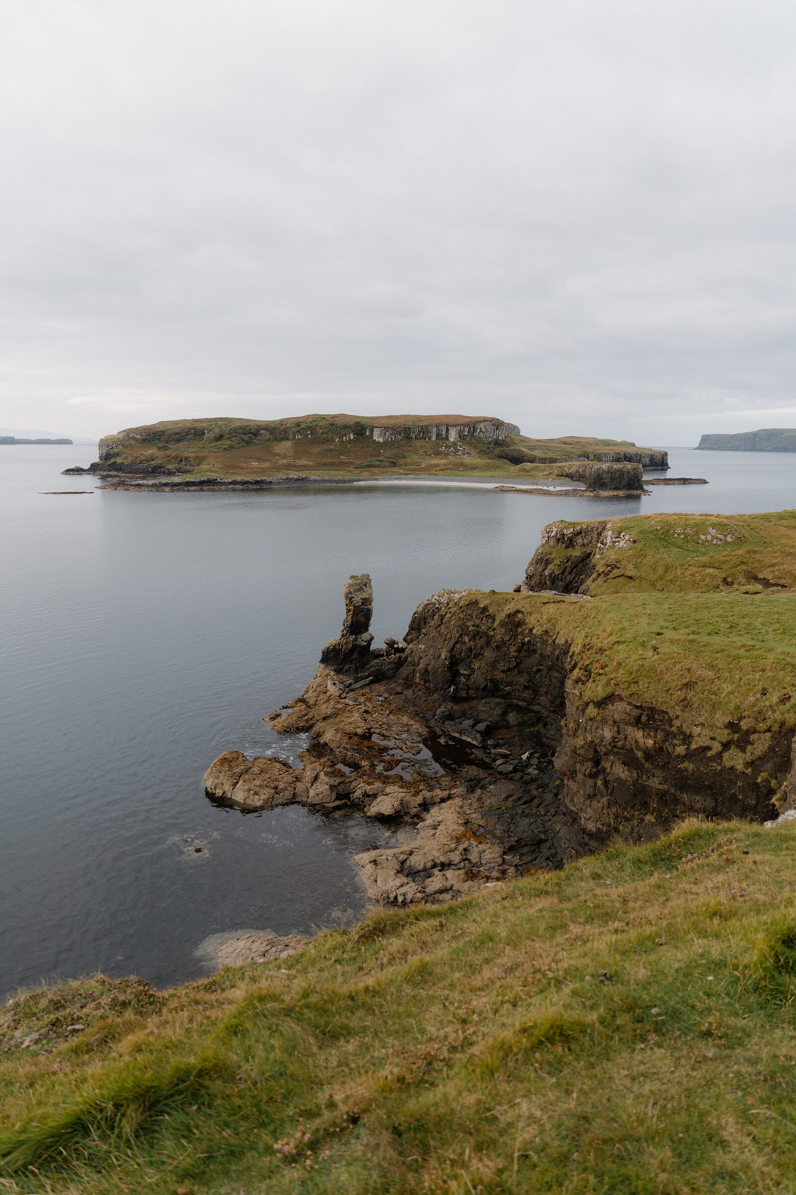 A rocky sea stack rises before a flat-topped island in calm water off the Isle of Skye.