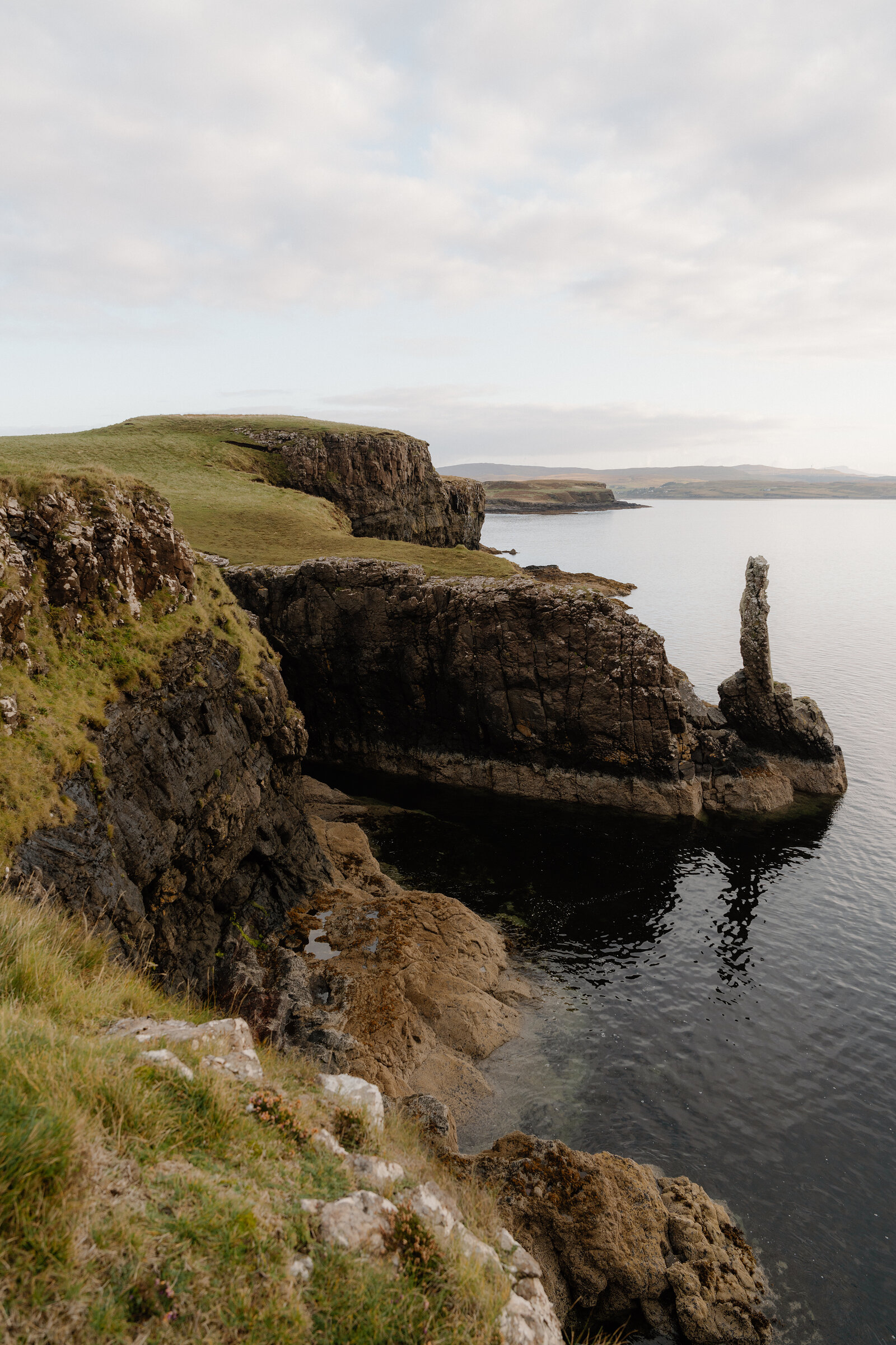 The grass-topped cliffs of Harlosh Point running out to the pillar of rock, calm water below.