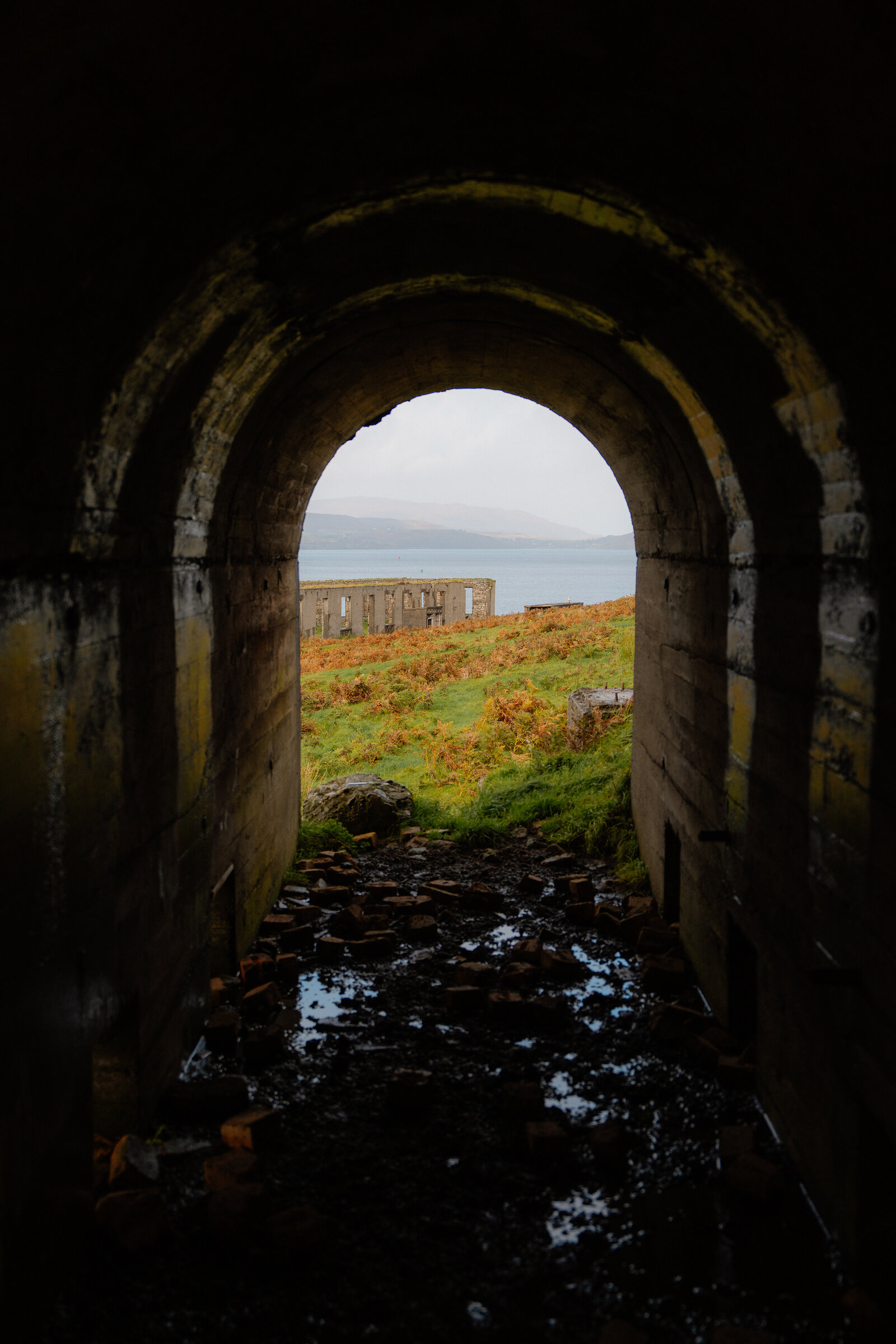 A stone archway frames a view of ruined industrial buildings and the sea on the Isle of Skye.