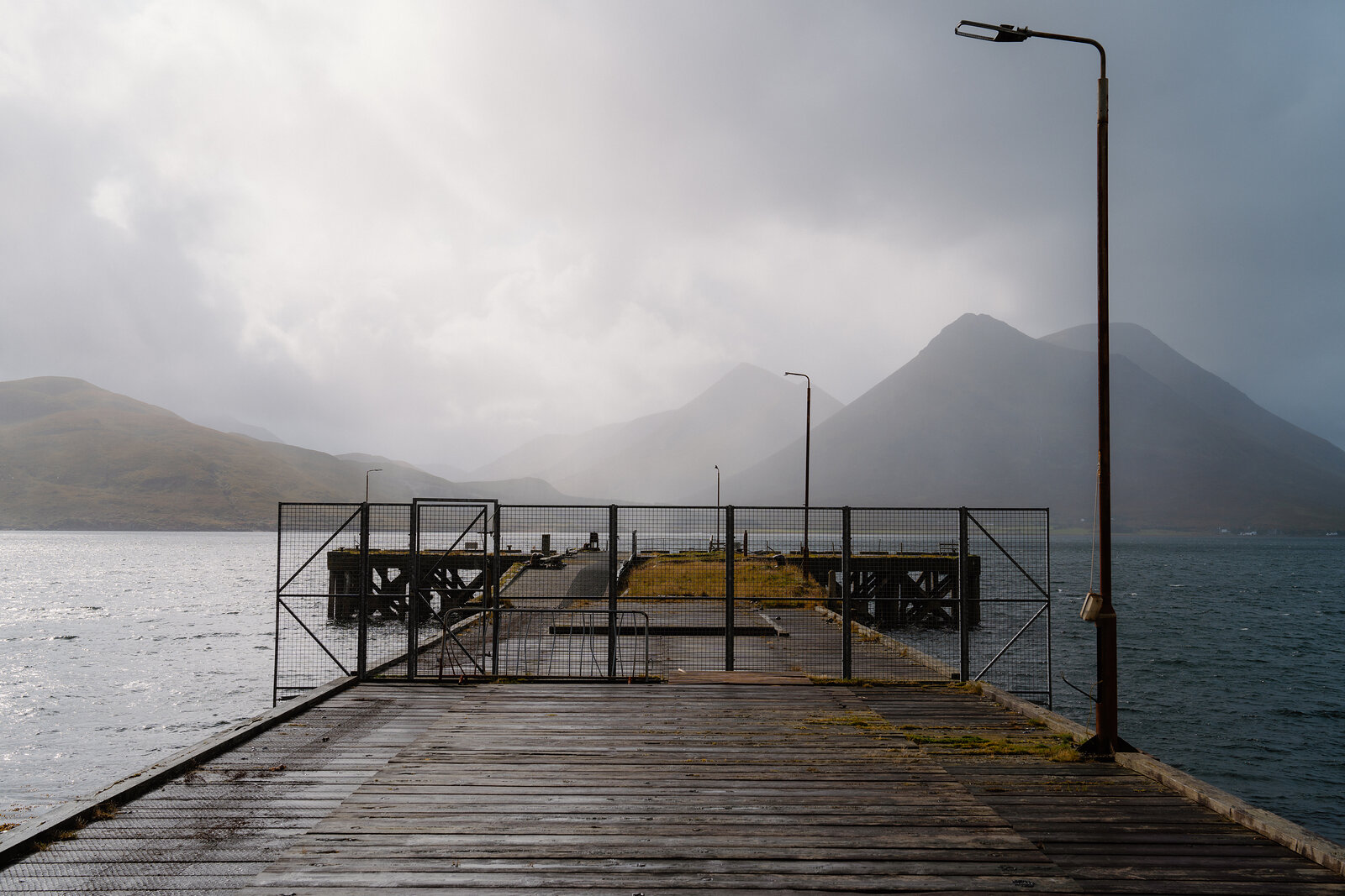 A wooden jetty with metal gates reaches into calm water beneath misty mountains on the Isle of Skye.