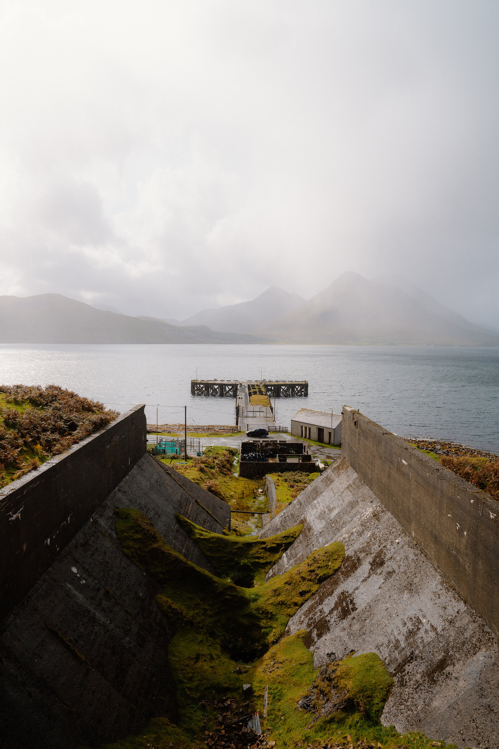 A metal jetty sits beside industrial ruins with misty mountains beyond on the Isle of Skye.