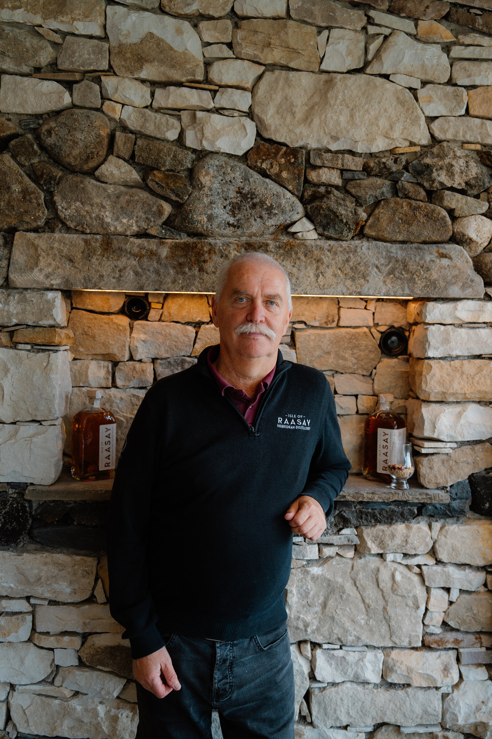 A man in a Raasay-branded fleece stands before a stone wall lined with whisky bottles at the Isle of Raasay Distillery.