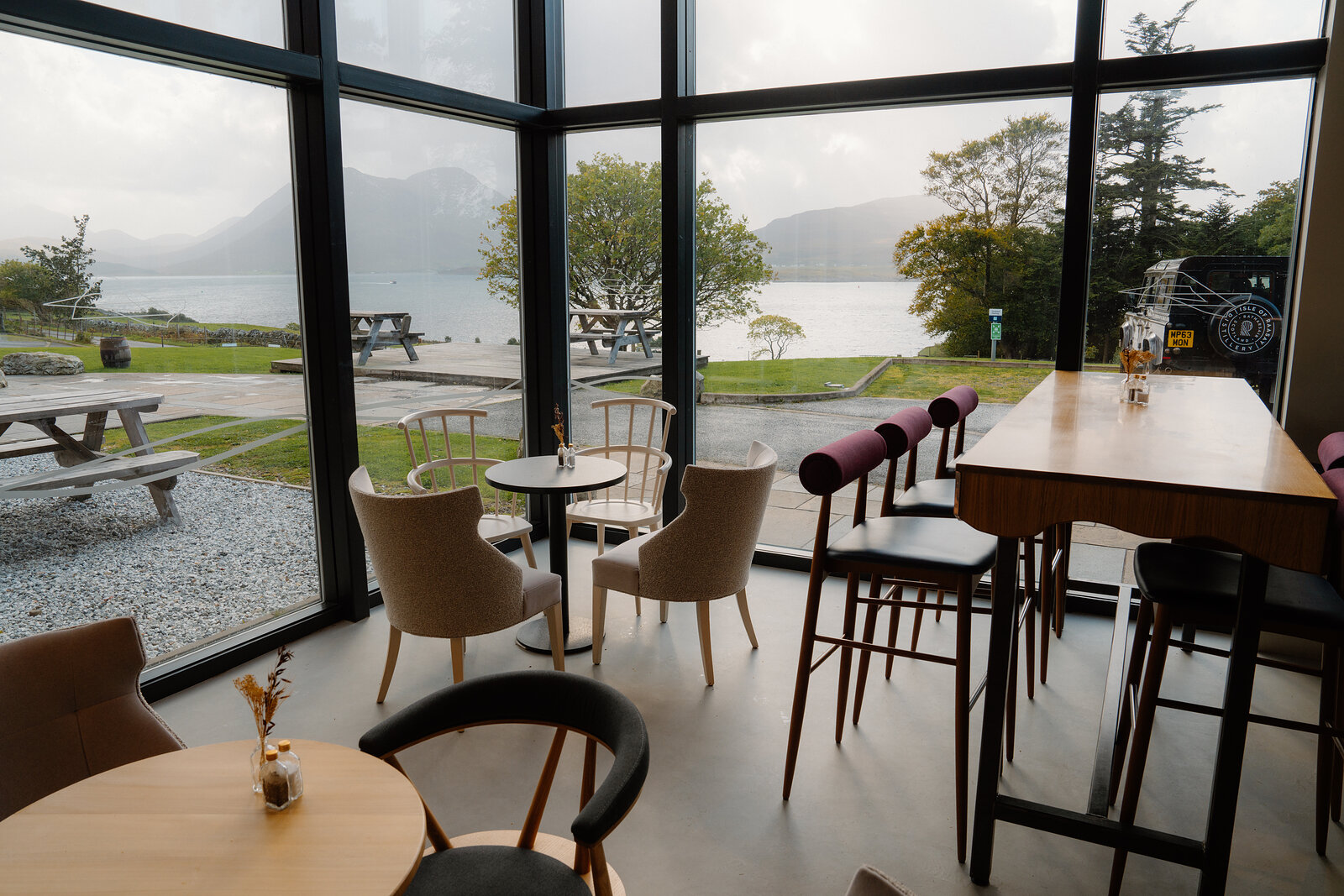 Cafe tables sit beneath large windows overlooking a rainy loch at the Isle of Raasay Distillery.