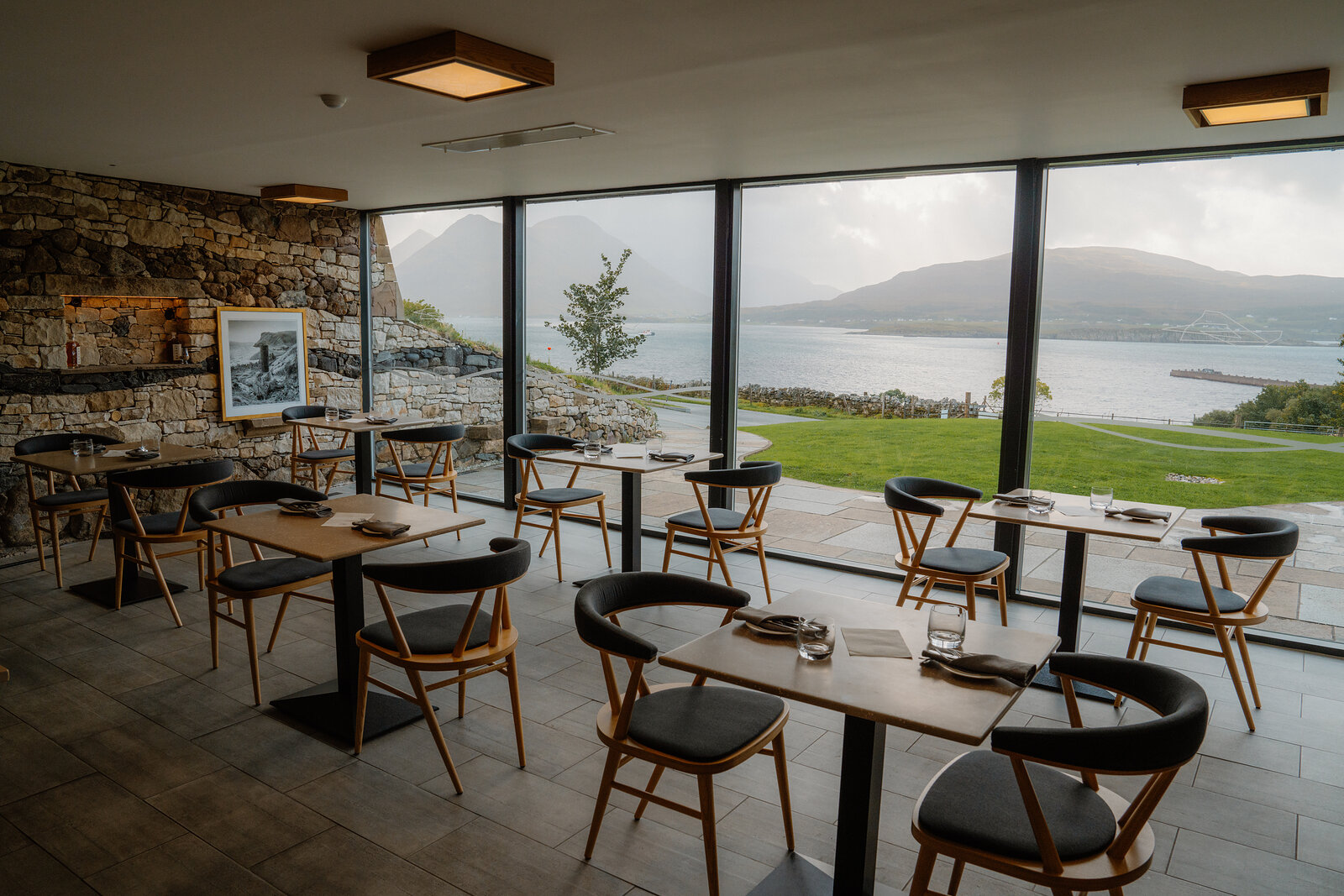 Tables are set for dining beside a stone wall and loch-view windows at the Isle of Raasay Distillery.